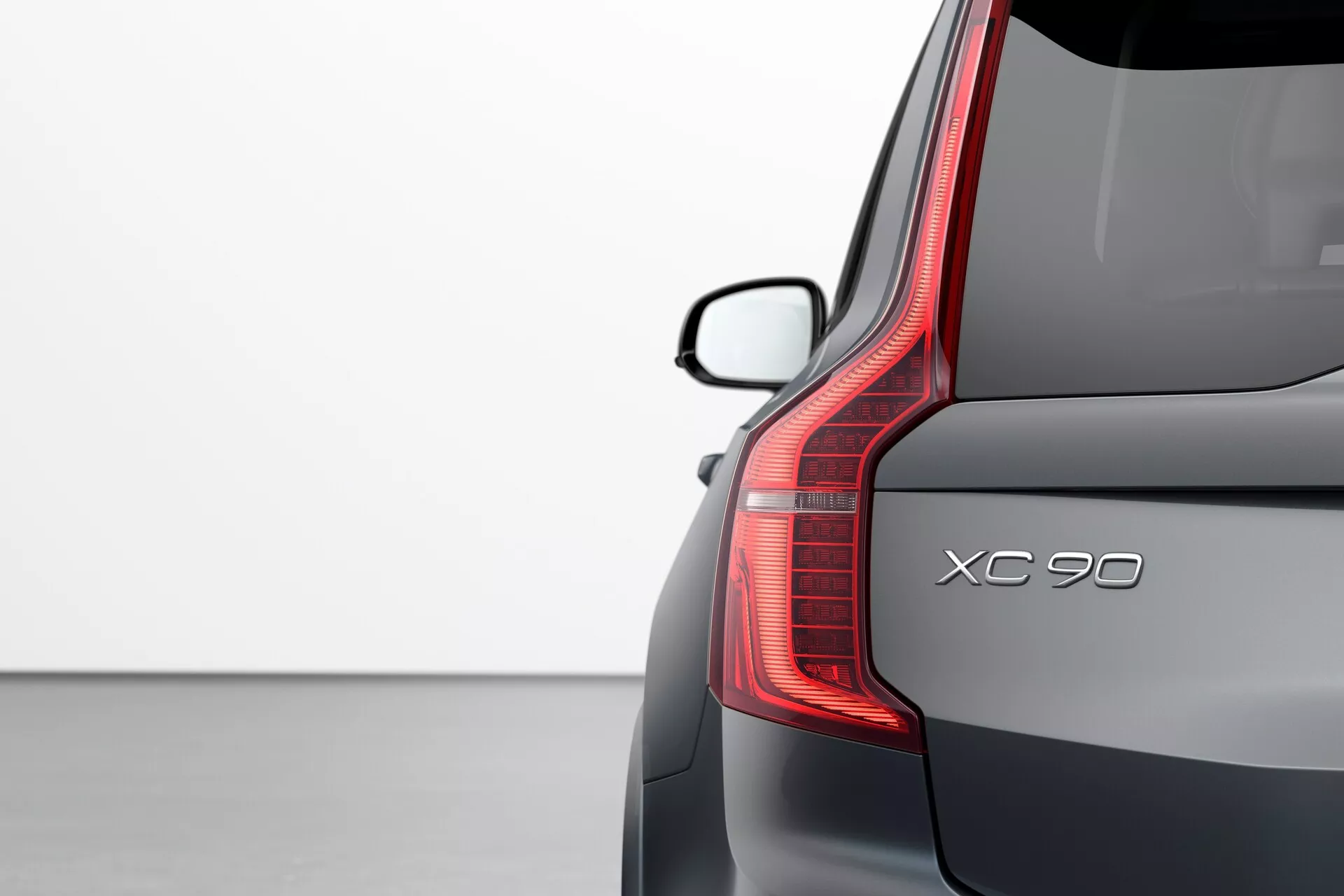 Vista ravvicinata dell'angolo posteriore di una Volvo XC90 2015 (2ª generazione) con evidenziati dettagli di design: fanale posteriore a LED verticale, nome del modello 'XC90', parabrezza posteriore elegante con spoiler e finitura della vernice metallica premium su sfondo chiaro.