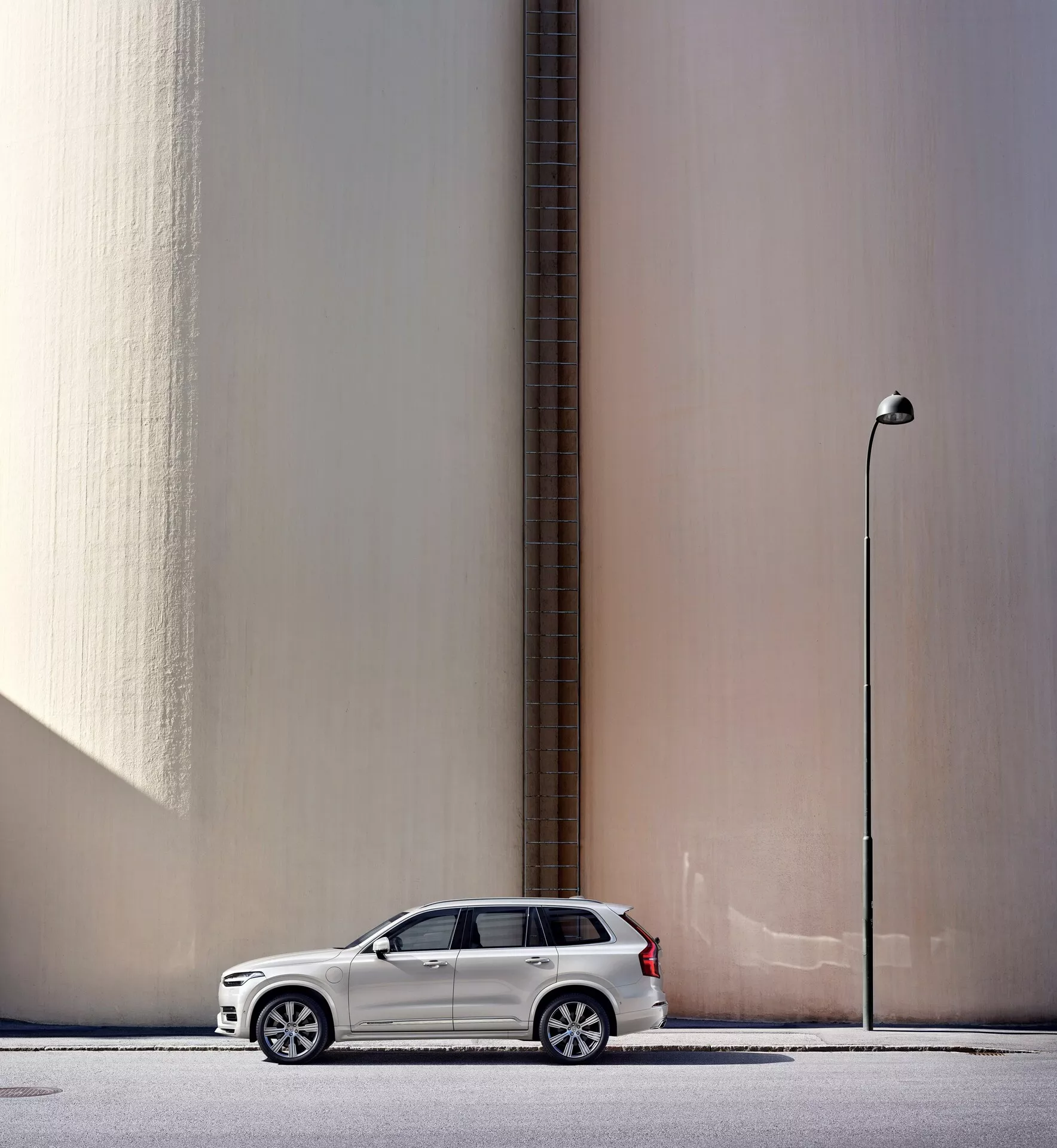 Visuale di profilo laterale di un SUV Volvo XC90 2015 bianco parcheggiato parallelamente a un muro beige alto e liscio con un tubo verticale, con uno sfondo urbano che include un lampione. L'auto, posizionata a sinistra del fotogramma con gli specchietti laterali spiegati, proietta un'ombra leggera che indica la luce naturale del giorno. L'immagine mette inoltre in risalto elementi di design moderno come cerchi in lega e fari LED, evidenziando caratteristiche fondamentali dell'auto.