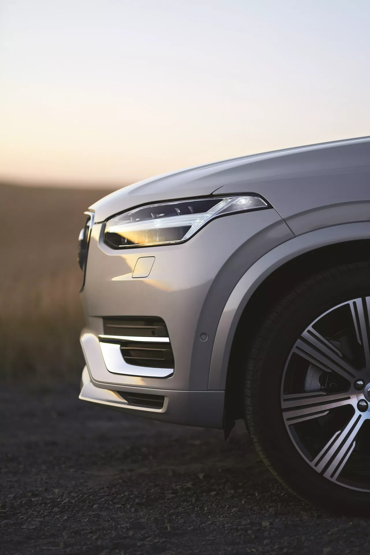 Vista del quarto anteriore sinistro del SUV di lusso Volvo XC90 seconda generazione 2015 all'alba, luci diurne a LED illuminate, vernice metallizzata che riflette la luce ambientale, elegante ruota in lega di grandi dimensioni, contorni intricati del cofano, design elegante del faro a LED, paraurti anteriore integrato con prese d'aria, sensori di assistenza alla guida potenzialmente visibili, profondità di campo ridotta con sfondo sfocato.