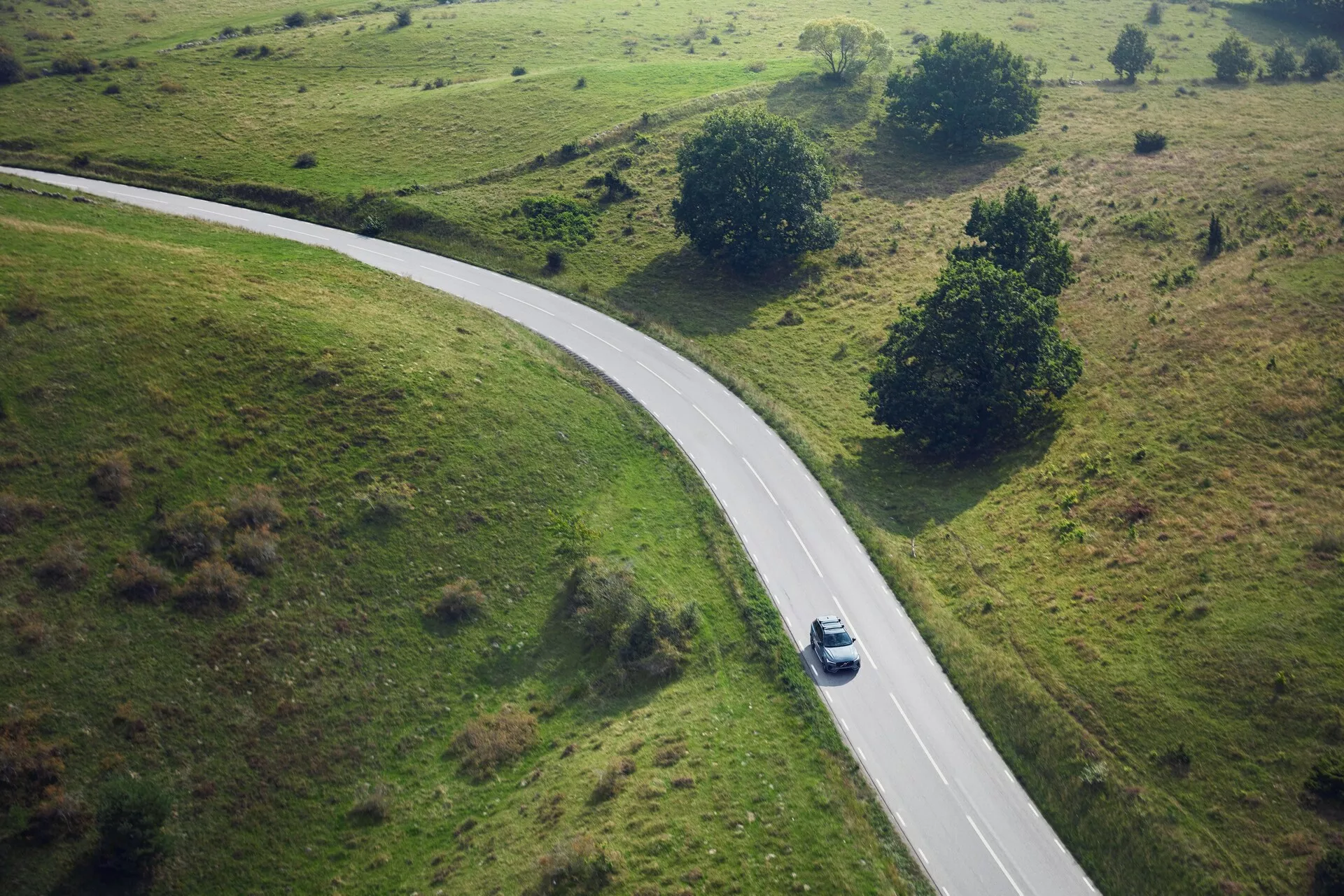 Vista aerea di una Volvo XC90 2015 argento o grigio chiaro che guida lungo una strada rurale curva circondata da campi d'erba con piccoli alberi e arbusti. L'auto, catturata da una prospettiva dall'alto, è posizionata a metà della curva al centro a destra dell'inquadratura, proiettando un'ombra che suggerisce il sole di mezzogiorno.