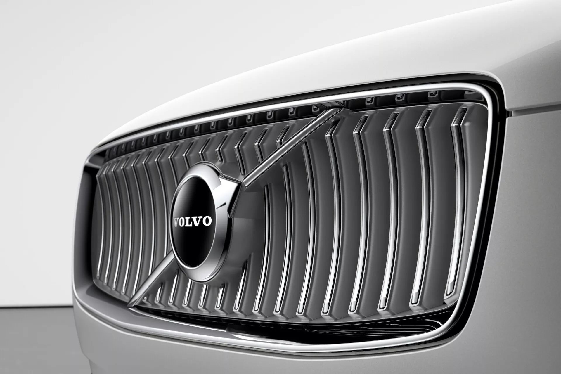 Vista ravvicinata della griglia frontale di una Volvo XC90 2015 (2a Generazione) da un angolo a tre quarti anteriore, con il prominente emblema Volvo posizionato al centro delle lamelle cromate verticali, sotto una luce soffusa che evidenzia la vernice chiara dell'auto, probabilmente bianca o grigio chiaro, e il design concavo ed elegante della griglia.