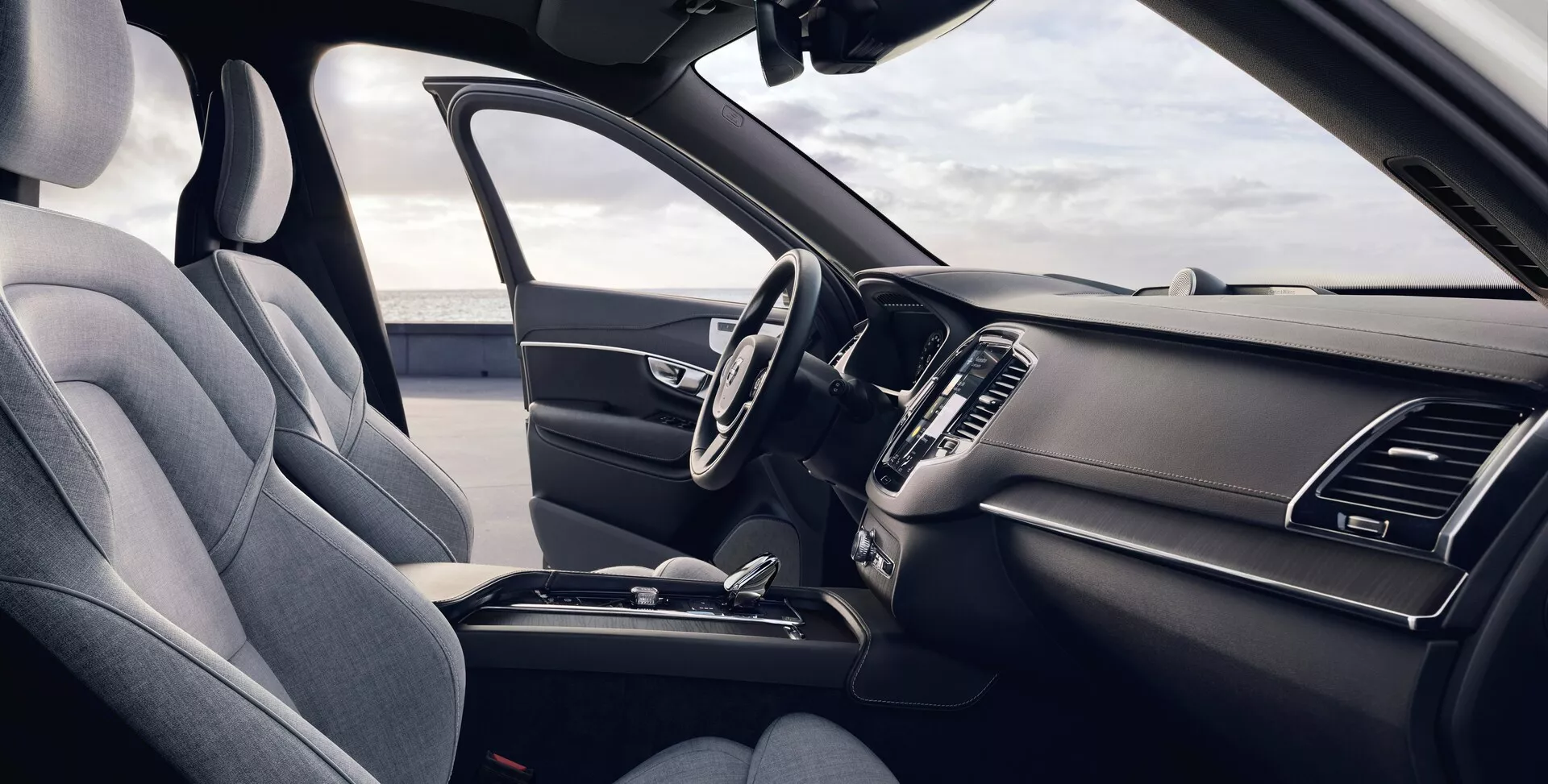 Vista interna di una Volvo XC90 2015 (2ª generazione) dalla prospettiva del passeggero anteriore, che mostra il sedile del conducente ergonomico rivestito in tessuto premium scuro o pelle sintetica, volante multifunzione con comandi integrati, cruscotto elegante con sistema di infotainment touch spento, prese d'aria integrate, controlli climatici minimali, console centrale con vano portaoggetti, portabicchieri e leva del cambio, tutto evidenziato in uno schema di colori scuri unificati con luce naturale che illumina l'auto.