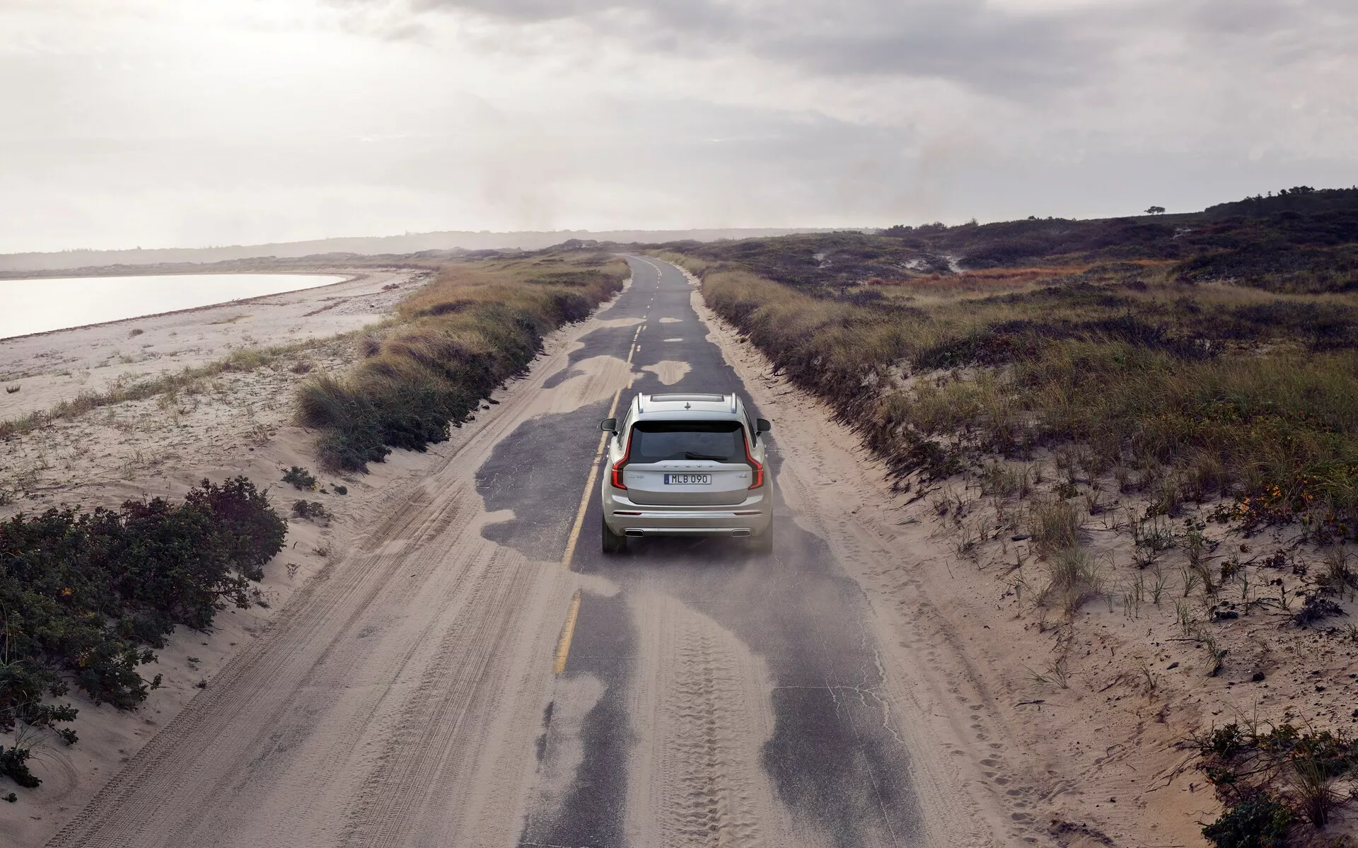 Vista aerea di un SUV Volvo XC90 2015 (2ª generazione) che guida su una strada sterrata stretta fiancheggiata da vegetazione bassa. Il veicolo moderno, dotato di antenna e barre sul tetto, è leggermente decentrato a sinistra e solleva polvere, suggerendo movimento e la superficie ruvida della strada. L'illuminazione nuvolosa illumina uniformemente la scena, contribuendo a una sensazione di isolamento. Le caratteristiche visibili includono luci posteriori, un tergicristallo posteriore e forse sensori di parcheggio.