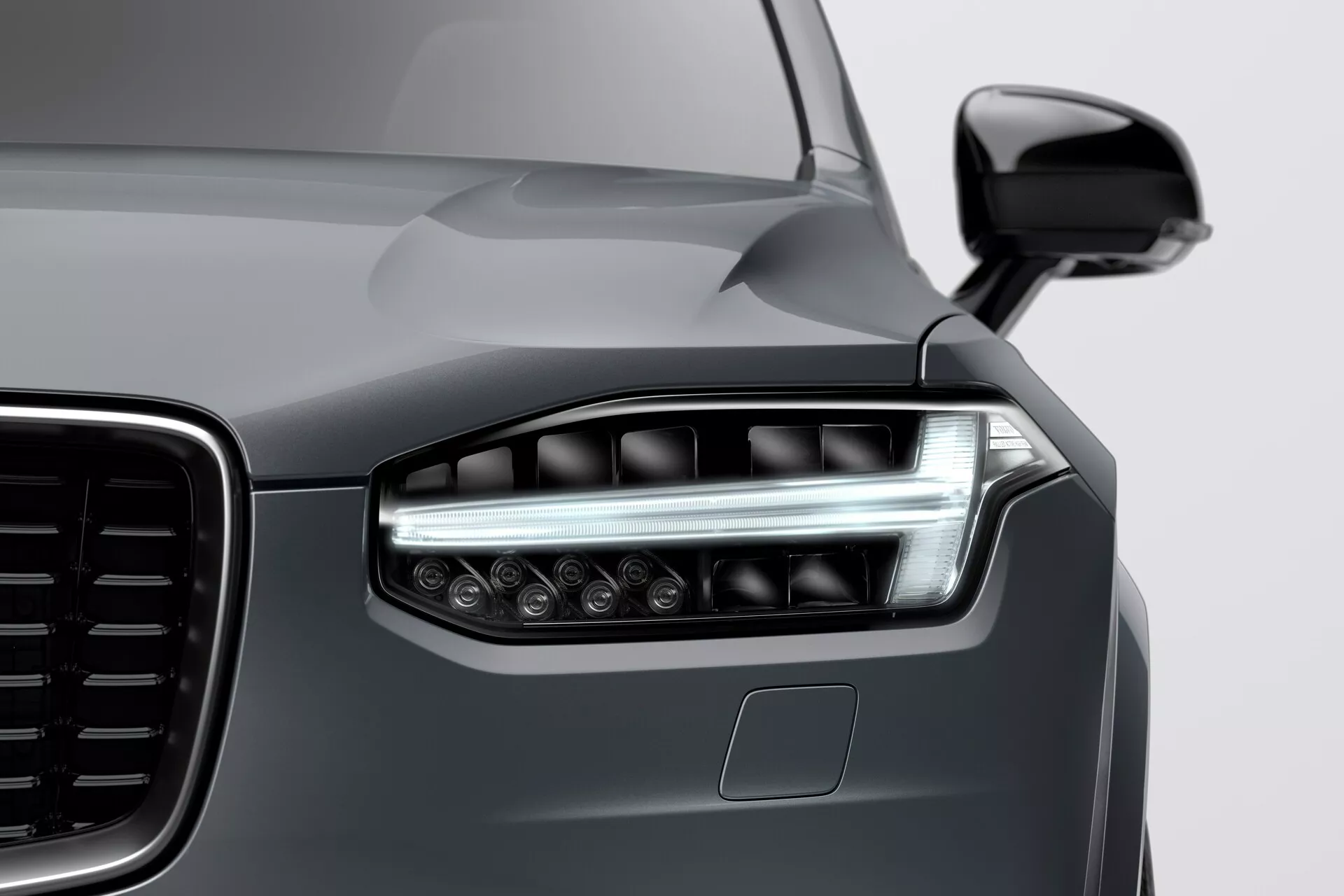 Vista ravvicinata dell'angolo anteriore sinistro di una Volvo XC90 2015 (2a generazione) che mostra un design moderno dei fari a LED con luci diurne angolari, una vista parziale della grande griglia con motivo verticale, vernice metallica grigio scuro lucida e sensore di parcheggio sul paraurti, scattata da un’angolazione bassa da destra a sinistra.