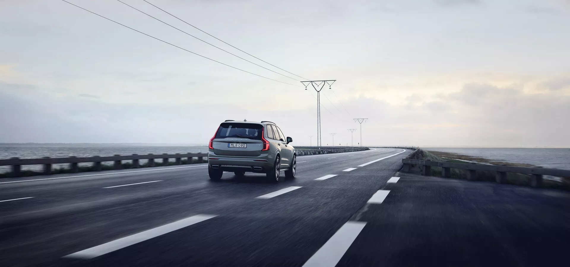 Vista posteriore tre quarti di un SUV in movimento su una carreggiata a doppia corsia con sistema di guida a destra. Il veicolo, posizionato nella corsia di destra, è probabilmente un SUV data l'altezza da terra rialzata e le proporzioni squadrate. Le caratteristiche visibili includono fanali posteriori, vista parziale del lato destro con porta passeggeri posteriore e pneumatico, portapacchi, ruote in movimento, paraurti posteriore e riflettori. La strada è umida con riflessi che indicano una finitura lucida dell'auto. La scena circostante consiste in un cielo nuvoloso, paesaggio cupo con pali elettrici, suggerendo un ambiente recentemente piovoso. Lo scatto cattura sia l'auto che il fondo in un'ampia inquadratura con profondità di campo, evocando temi di tecnologia, viaggi e attività all'aperto con illuminazione naturale e soffusa.
