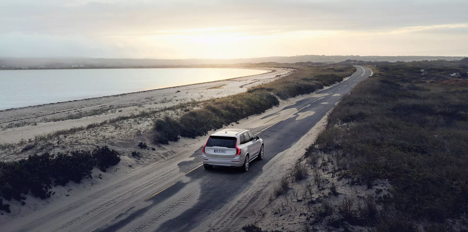 Scatto aereo di un SUV Volvo XC90 2015 (2ª generazione) bianco o di colore chiaro che guida su una strada sterrata tortuosa parallela a un corpo d'acqua, possibilmente un fiume, con un paesaggio costiero sulla destra. La scena è ambientata all'alba o al tramonto con una luce naturale soffusa che proietta lunghe ombre, accentuando i dettagli dell'auto e del suo ambiente. L'auto è leggermente a destra del centro nel terzo inferiore dell'immagine, mostrando la parte posteriore, inclusi il portellone e il parabrezza posteriore. L'immagine enfatizza un senso di avventura e viaggio in un ambiente naturale pittoresco.