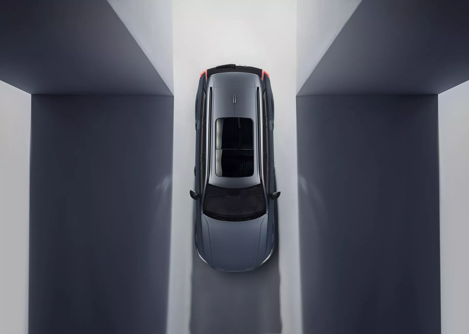 Vista aerea di una Volvo XC90 2015 (2ª generazione) che mostra una composizione simmetrica e riflessi ambientali. La posizione centrale in un ambiente interno dai toni neutri mette in evidenza il tetto dell'auto con tetto apribile o tetto panoramico in vetro. Le parti visibili includono il cofano, il tetto, il bagagliaio e la parte superiore delle portiere, enfatizzando linee eleganti e curve che suggeriscono un design lussuoso focalizzato sull'aerodinamica e un'illuminazione interna spaziosa.