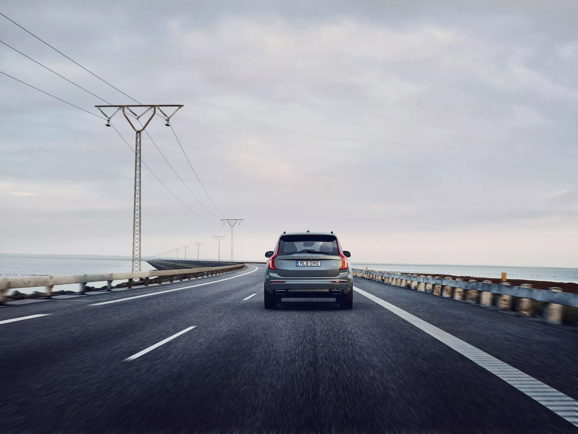 Vista posteriore di una Volvo XC90 del 2015 (2ª generazione) che viaggia su una strada costiera a due corsie. La foto è scattata da un'angolazione bassa, con una prospettiva centrata, catturando il portellone, il parabrezza posteriore, i fanali posteriori, il paraurti posteriore e i badge di marca e modello. Il veicolo è il soggetto principale, con un orizzonte oceanico leggermente sfocato e torri elettriche che si estendono in lontananza sotto un cielo grigio e nuvoloso. Il numero di targa del veicolo è visibile e l'illuminazione suggerisce la prima mattina o il tardo pomeriggio, con luce diffusa.