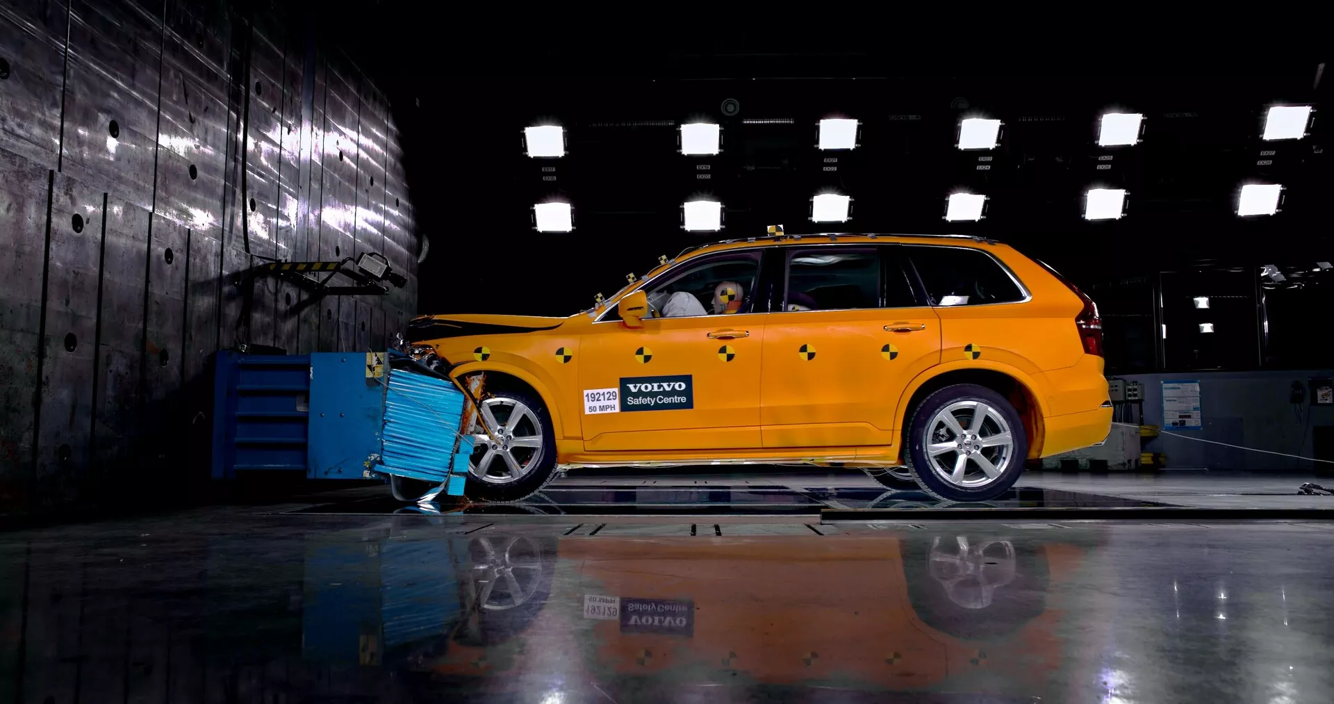 Vista di profilo laterale di una Volvo XC90 2015 (2a generazione) gialla brillante in un ambiente industriale, che mostra il suo design station wagon con griglia anteriore, fari e ruote in lega visibili. L'auto è collegata a un'attrezzatura diagnostica o di ricarica blu e un manichino all'interno suggerisce una dimostrazione di test di sicurezza o crash test. Il pavimento lucidato riflette il telaio e le ruote del lato opposto.