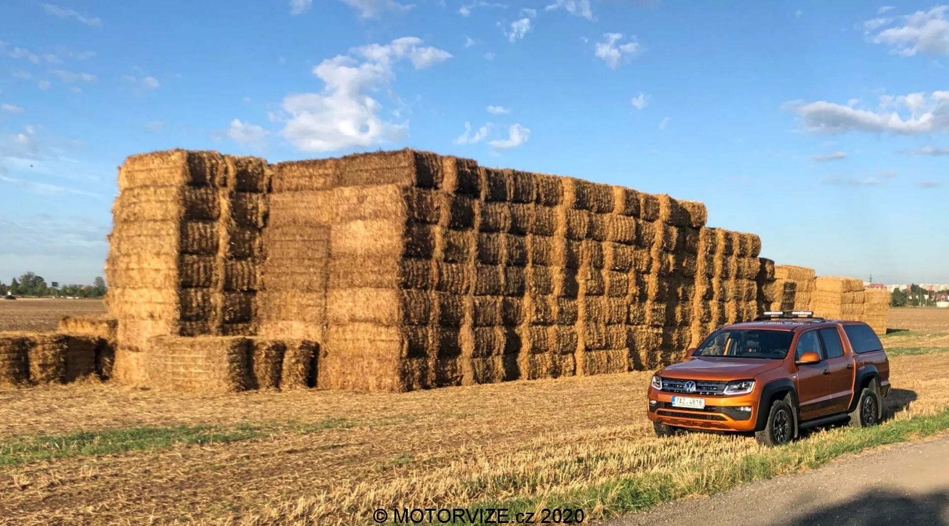 Un Volkswagen Amarok 2016 parcheggiato davanti a grandi balle di fieno in un contesto rurale. La prospettiva frontale di tre quarti cattura la griglia, i fari, il paraurti anteriore, il cofano, le ruote, gli specchietti laterali e le porte dell'auto. La griglia a listelli orizzontali prominente e i fari angolari acuti migliorano l'aspetto contemporaneo del veicolo. I finestrini colorati, i binari sul tetto e i rivestimenti protettivi intorno alle porte suggeriscono versatilità e capacità robusta. Il veicolo è situato su uno sfondo di balle di fieno, aggiungendo contesto al suo utilizzo per la guida in campagna o agricola. La luce del mattino o del tardo pomeriggio proietta ombre calde e migliora i dettagli del colore e del design dell'auto. Con filigrana 'MOTORVIZE.cz 2020'.