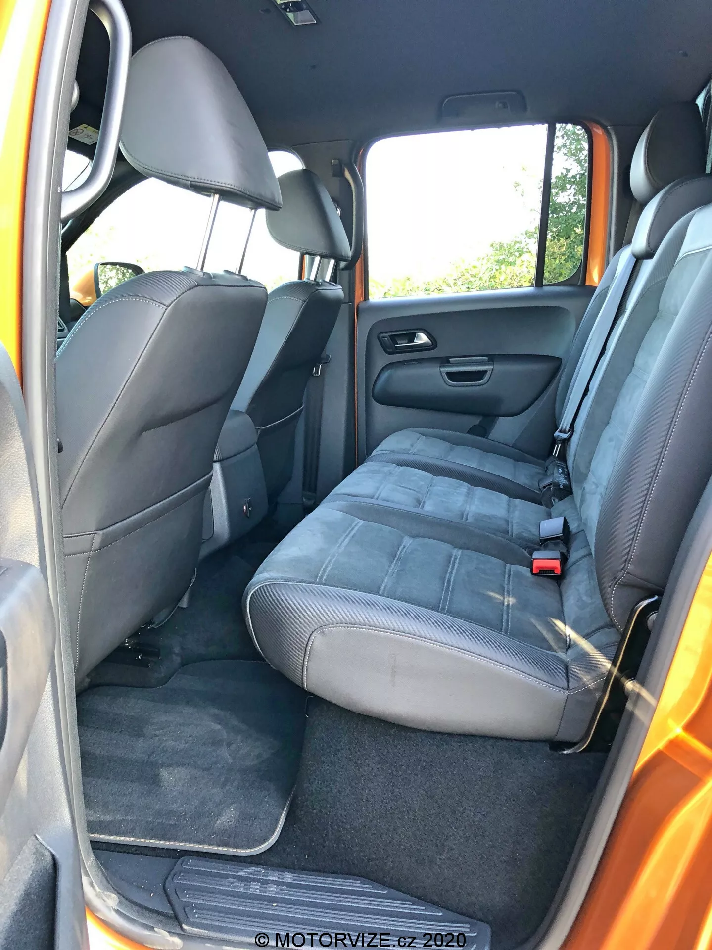 Vista interna del VW-Nutzfahrzeuge Amarok 2016 da un angolo della porta del passeggero posteriore; mostra un sedile posteriore a panca adatto per tre passeggeri con rivestimento in tessuto, poggiatesta, cinture di sicurezza (cintura addominale al centro, cinture a tre punti sui lati), porta aperta che rivela il pannello con altoparlante, vano portaoggetti e controllo del finestrino elettrico; pavimento moquette e sedili posteriori pieghevoli; tasca di stivaggio sul retro del sedile passeggero anteriore; caratteristiche standard dell'auto senza elementi tecnologici rilevanti; luce naturale.