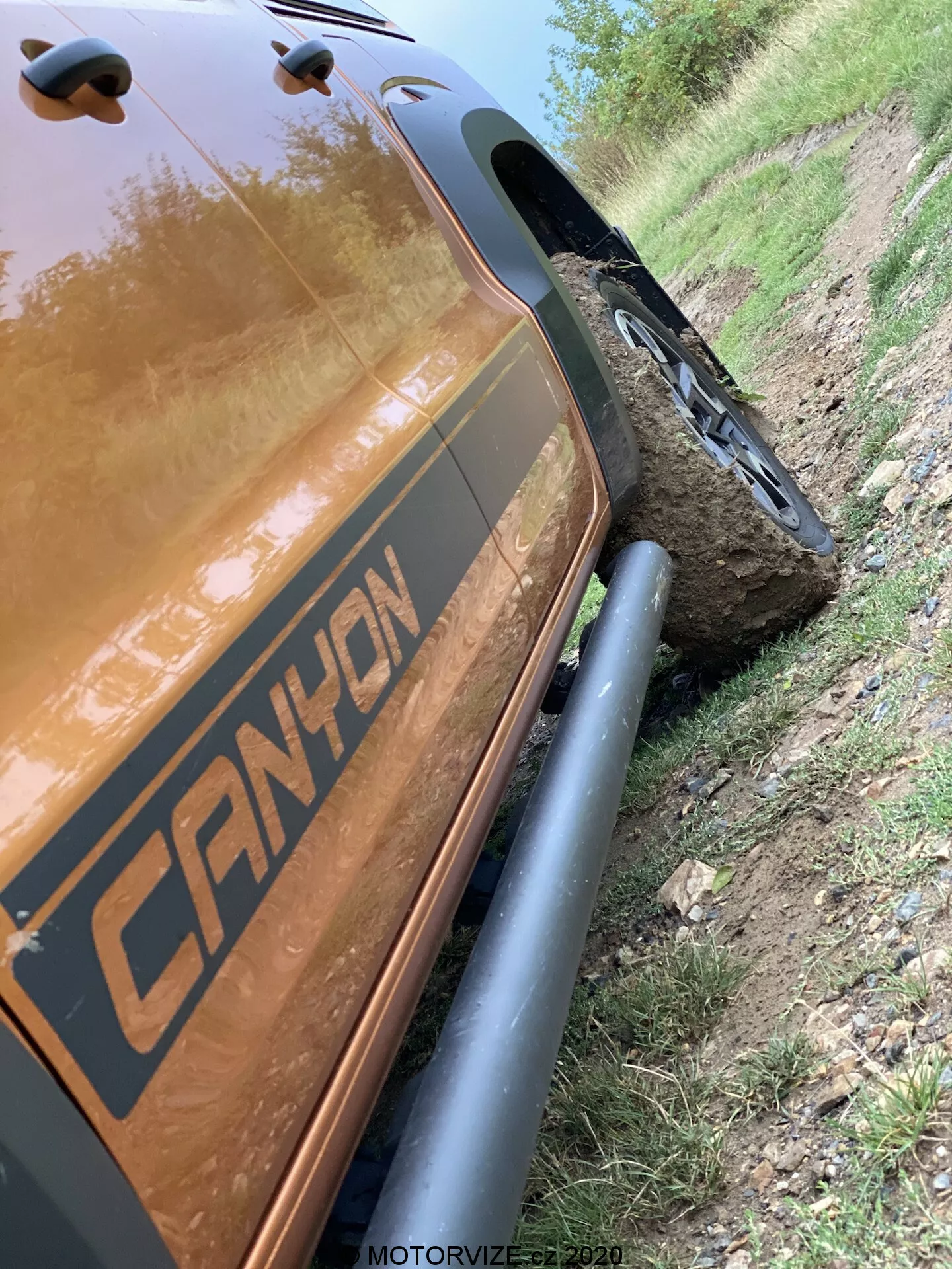 Questa immagine inclinata a 45 gradi mostra la sezione posteriore del lato passeggero di un VW-Nutzfahrzeuge Amarok 2016. Si concentra sui dettagli ravvicinati dall'arco della ruota posteriore alla sezione di carico posteriore, evidenziando robuste protezioni nere dei passaruota e pneumatici fuoristrada robusti. Il veicolo presenta una grafica con l'etichetta 'CANYON' ed è posizionato accanto a un sentiero polveroso con una ruota montata su una grande pietra, dimostrando le sue capacità fuoristrada. Il colore riflettente metallico marrone o rame della carrozzeria è evidente, catturando riflessi di luce. Il timestamp 'MOTORVIZE.cz 2020' suggerisce che è stato catturato per una pubblicazione di veicoli a motore.