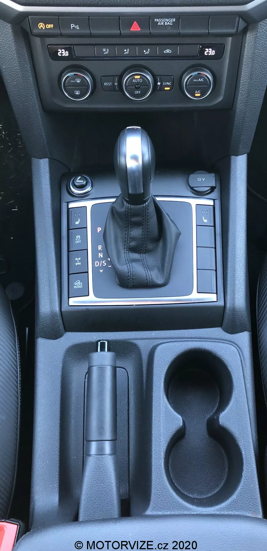 Questa immagine mostra la console centrale di un vw-nutzfahrzeuge amarok 2016, presa da una prospettiva aerea all'incirca dal punto di vista del conducente o del passeggero anteriore. La fotografia cattura l'interno dell'auto specifico per la leva del cambio, i controlli della console centrale e i vani portaoggetti. L'immagine include quadranti e pulsanti del sistema di controllo del clima, una leva del cambio rivestita in pelle su un pannello argentato che indica una trasmissione automatica, un controllo del freno di stazionamento elettronico, un vano portaoggetti, due portabicchieri e un ulteriore vano verso i sedili posteriori. I materiali visibili includono plastica, accenti metallici e pelle, riflettendo una combinazione di funzionalità e estetica.