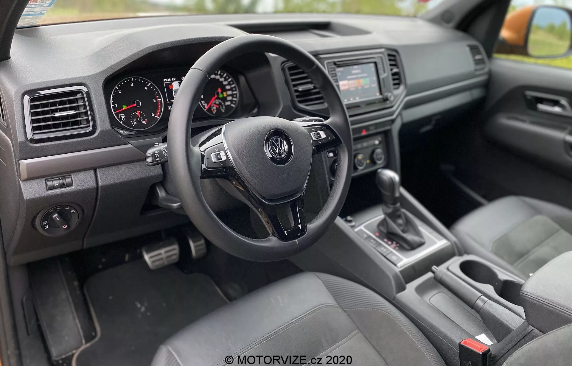 Vista interna del VW Nutzfahrzeuge Amarok 2016 dal lato passeggero, con guida a destra, volante a tre razze Volkswagen con pulsanti di controllo, quadro strumenti con tachimetro e contagiri analogici, console centrale con sistema audio e schermo di infotainment, leva del cambio in posizione 'P', sedili rivestiti in tessuto, cruscotto con bocchette d'aria e plastica testurizzata e pannello della portiera del conducente con comandi dei finestrini e regolazione degli specchietti.