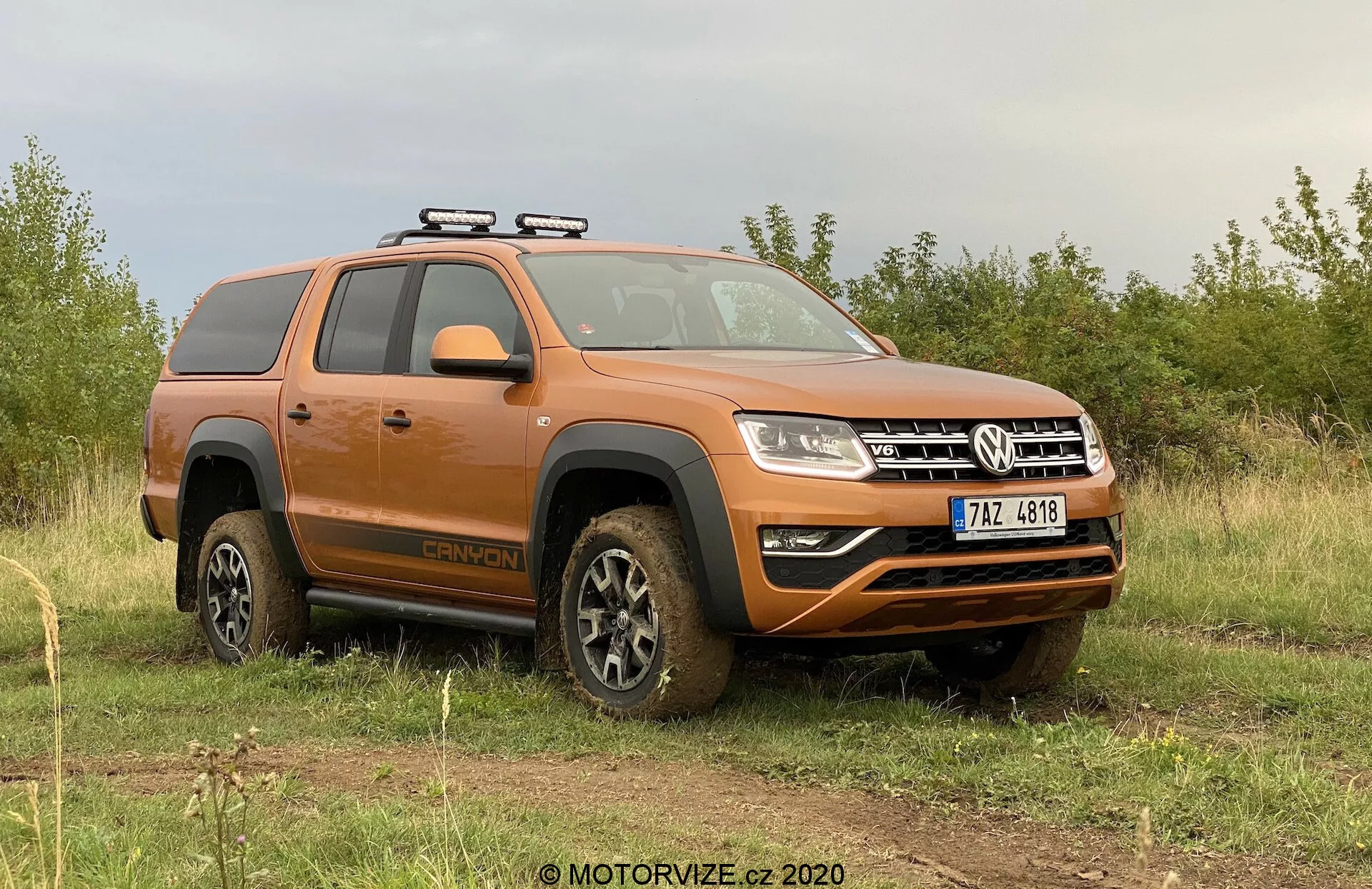 L'immagine mostra un pickup Volkswagen Amarok 2016 arancione parcheggiato su un terreno erboso irregolare con uno sfondo di cielo nuvoloso. Il prospetto laterale-frontale si concentra sul lato del conducente, mostrando il profilo del veicolo e una parte della parte anteriore. Le caratteristiche principali visibili includono un portapacchi, ruote coperte di fango che indicano attività fuoristrada recente, gradini laterali, configurazione a doppia cabina, design moderno dei fari e paraurti anteriore con fendinebbia e una piastra paramotore inferiore. Il veicolo emana un'estetica robusta e utilitaria, con elementi tecnologici moderni.