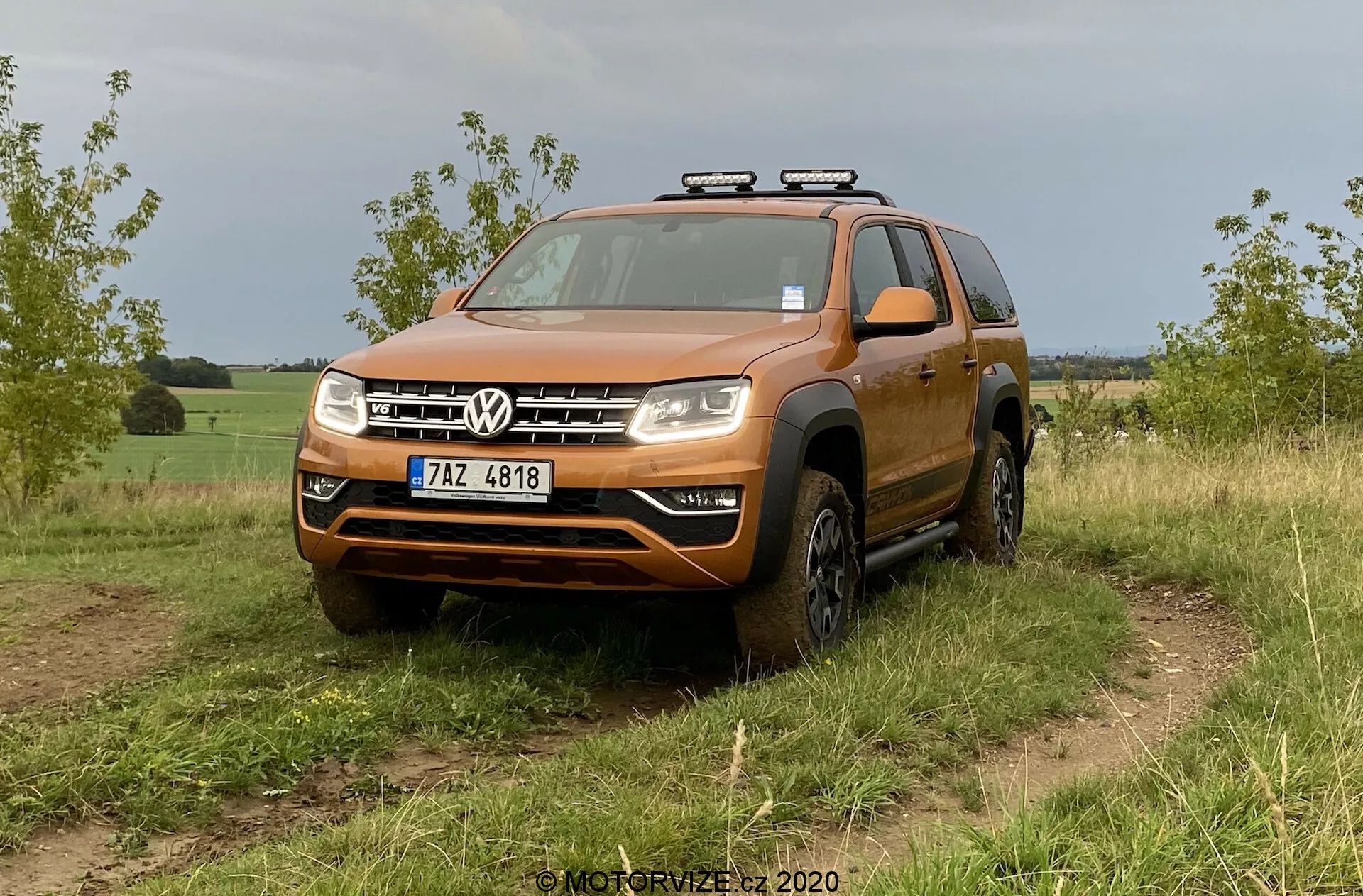 Un SUV Volkswagen arancione, nello specifico il VW Amarok 2016, è mostrato su un terreno fuoristrada erboso. L'inquadratura da un angolo frontale basso a medio enfatizza il design robusto, adatto per le attività fuoristrada. Il veicolo presenta una griglia frontale audace con due barre cromate che incorniciano il logo centrale Volkswagen, un paraurti considerevole con una probabile protezione sottoscocca, fari compositi chiari con potenziali lampadine LED o alogene e archi delle ruote svasati che ospitano pneumatici fuoristrada. Le luci ausiliarie sono montate sul tetto, suggerendo una funzionalità aggiuntiva per la guida notturna. I dettagli visibili includono il parabrezza, le barre sul tetto, gli specchietti laterali con indicatori di direzione integrati e la targa anteriore. Il veicolo appare pulito con un po' di sporco sui lati inferiori e sulle ruote, in linea con l'uso fuoristrada. Lo sfondo suggerisce un'ambientazione rurale o semi-rurale.