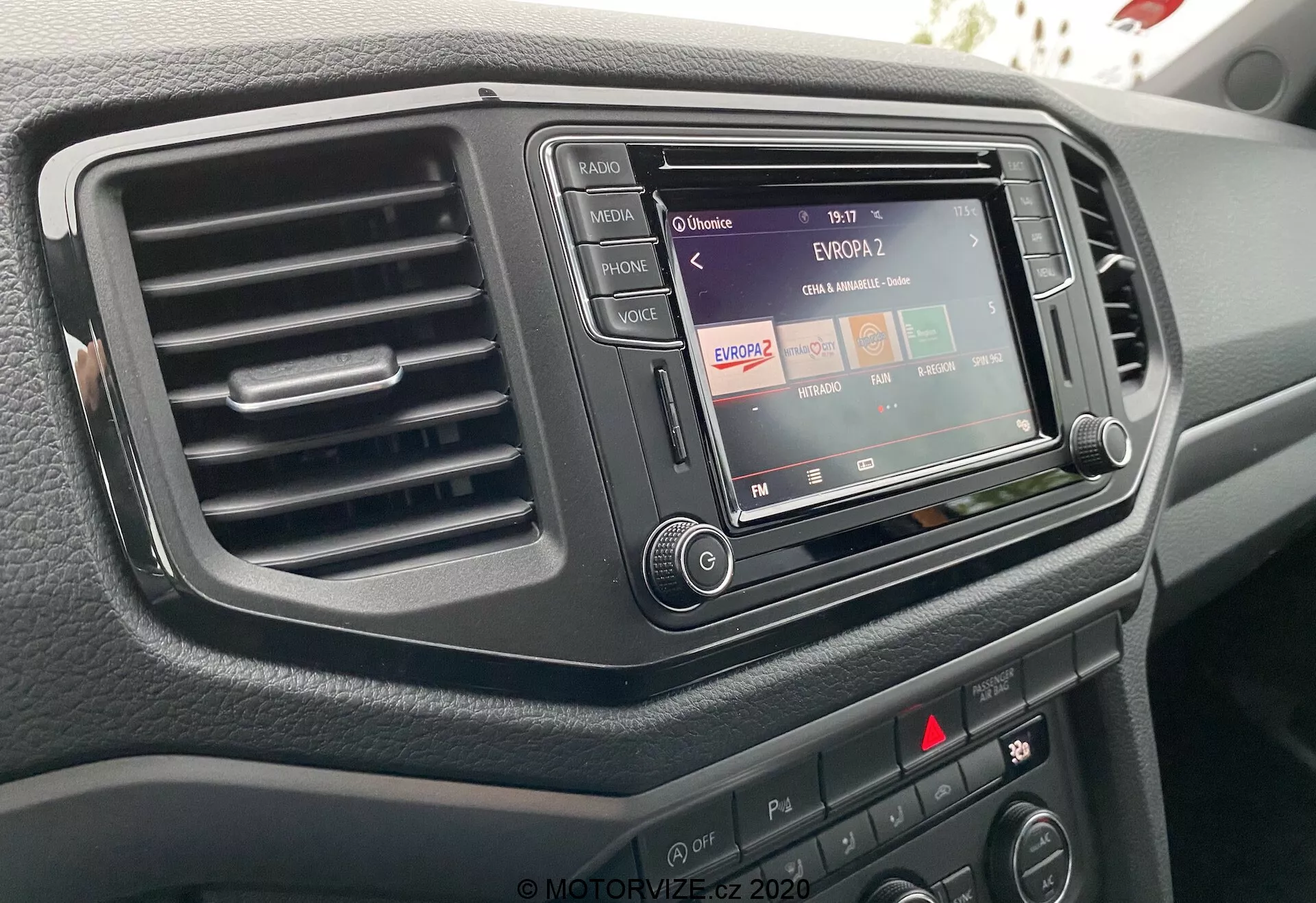 Primo piano della console centrale di un VW Nutzfahrzeuge Amarok 2016 dalla prospettiva del conducente, mettendo in evidenza il sistema di infotainment con funzionalità radio, una sola bocchetta d'aria a sinistra e vari pulsanti e manopole di controllo. Il sistema di infotainment è incastonato in un rivestimento scuro ed elegante e la superficie del cruscotto ha una finitura testurizzata.