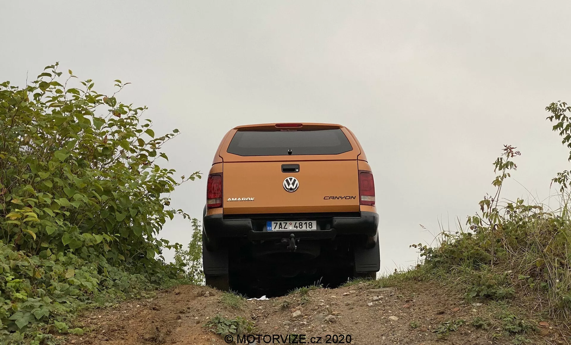 Vista posteriore di un crossover o SUV Volkswagen arancione da un'angolazione leggermente bassa, catturata sulla cresta di una piccola collina. Sono chiaramente visibili gli elementi di design posteriore del veicolo come il portellone, il finestrino posteriore, i fanali posteriori, i badge di marca e modello che leggono 'Volkswagen' e 'CROSSOVER' e il paraurti posteriore. La targa è visibile, ma non vengono forniti dettagli specifici. Il portellone presenta una piega prominente e linee di design, con una possibile maniglia o recessione sotto la targa di immatricolazione. I fanali posteriori si avvolgono sui lati e includono indicatori, luci di retromarcia e luci di arresto. La parte inferiore nera del paraurti posteriore suggerisce una finitura protettiva per l'uso fuoristrada. Il veicolo è incorniciato da vegetazione, indicando un ambiente rurale o fuoristrada. Il terreno è di terra naturale, coerente con le capacità fuoristrada del veicolo. Nell'angolo in basso a destra c'è una filigrana che recita 'MOTORVIZE.cz ©2020'.