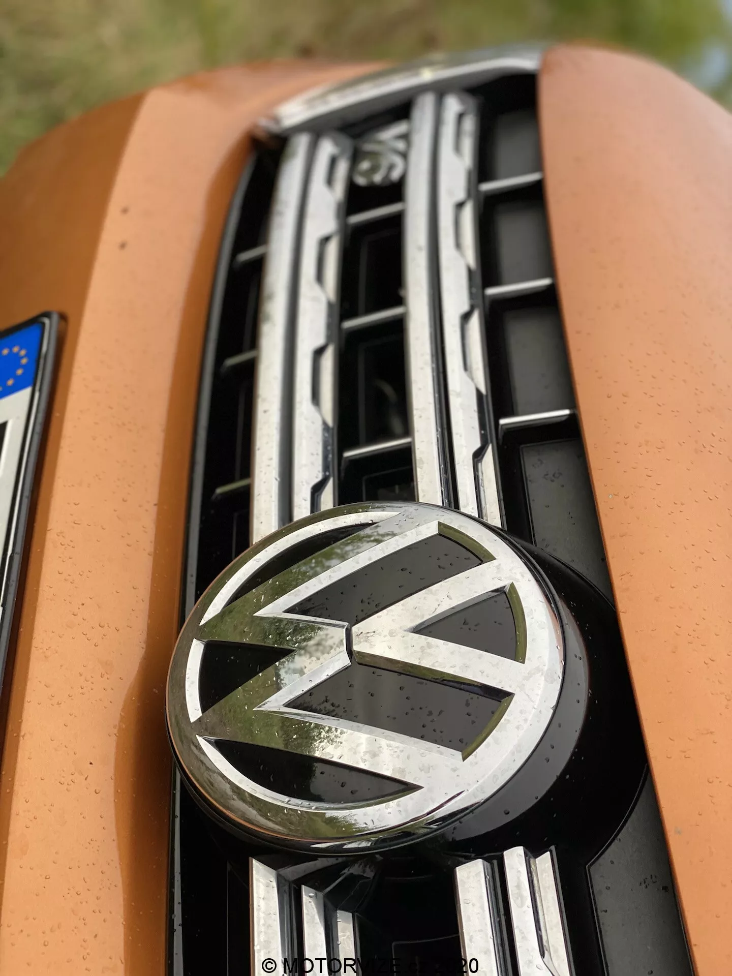 Primo piano dell'area della targhetta frontale della griglia di un veicolo Volkswagen. L'immagine si concentra sul logo VW incorniciato da lamelle orizzontali cromate, fotografato da una prospettiva bassa e leggermente frontale con una profondità di campo ridotta. Lo sfondo sfocato enfatizza il logo iconico Volkswagen e il design del frontale della vettura, evidenziando la vernice arancione sul cofano.