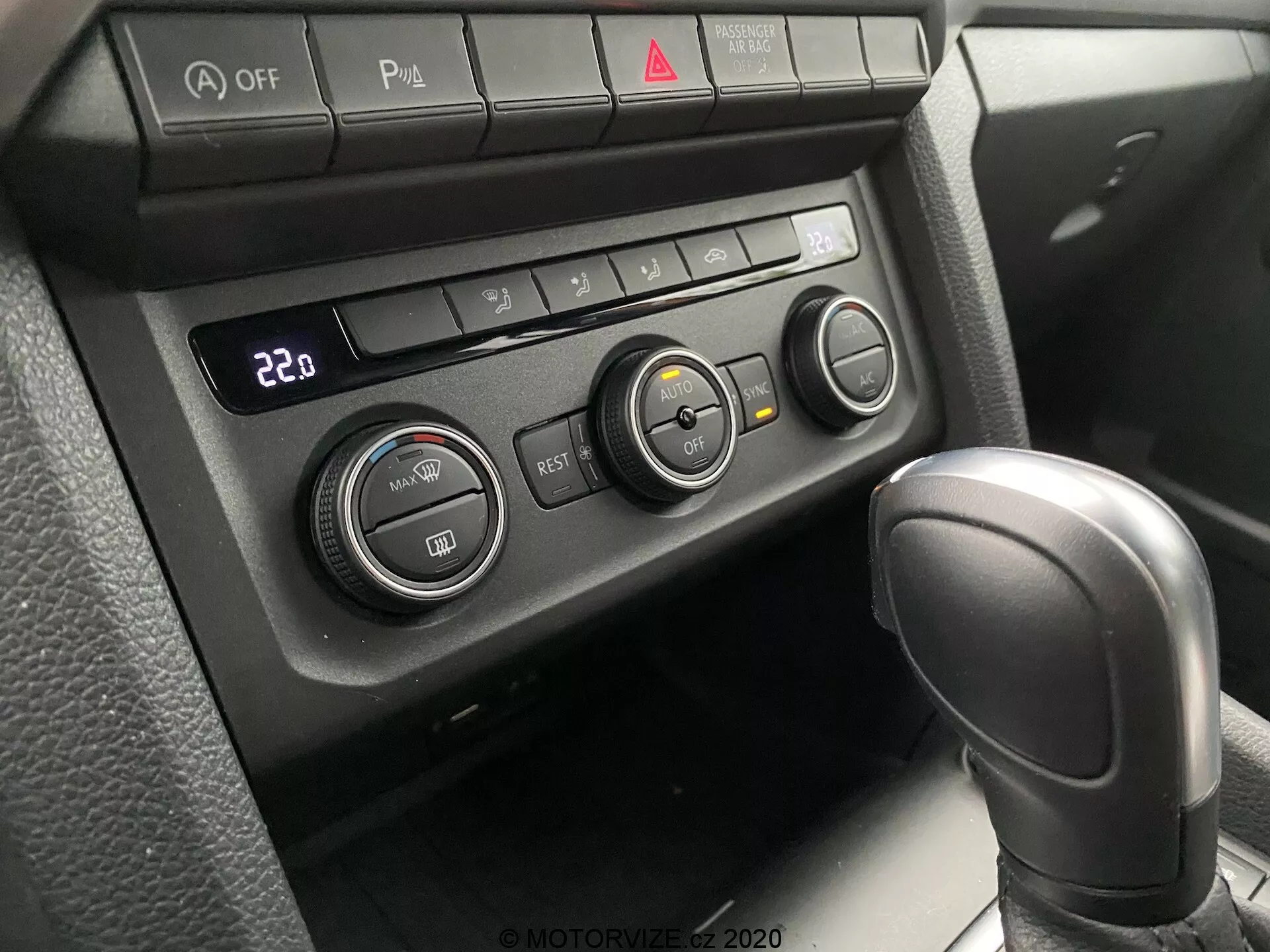Vista ravvicinata del sistema di controllo del clima e della parziale leva del cambio del VW Nutzfahrzeuge Amarok 2016. Prospettiva del passeggero o del conducente che guarda verso il basso alla console centrale. Presenta una leva del cambio automatica con texture in pelle in 'Park' e un pannello di controllo del clima impostato a 22°C con tre manopole rotanti e vari pulsanti funzionali. I pulsanti includono disattivazione Auto Stop/Start, interruttore sensori di parcheggio, volante riscaldato e sedile riscaldato, insieme a un pulsante della luce di emergenza rosso centrale. L'illuminazione morbida e diffusa suggerisce un interno ben ombreggiato.