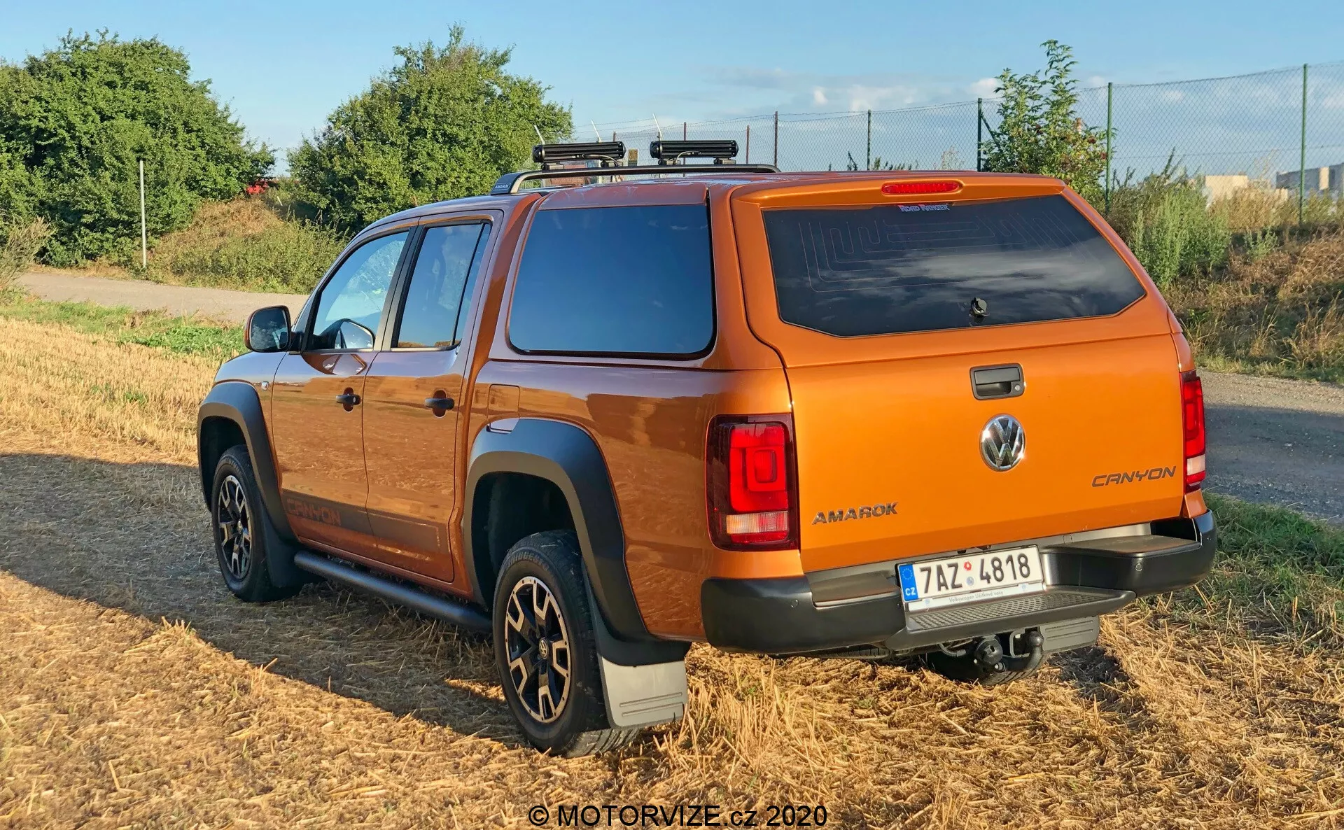 Vista posteriore tre quarti di un pickup Volkswagen Amarok 2016 arancione con tetto rigido, sistema di portapacchi e cerchi in lega multiraggi. Il veicolo è parcheggiato su un'area erbosa con luce naturale soffusa, mostrando il badge Volkswagen, il nome del modello 'AMAROK' e la designazione 'CANYON', e luci posteriori moderne. Le pedane laterali scure e la configurazione della cabina doppia evidenziano la sua funzionalità sia per il trasporto che per i passeggeri.