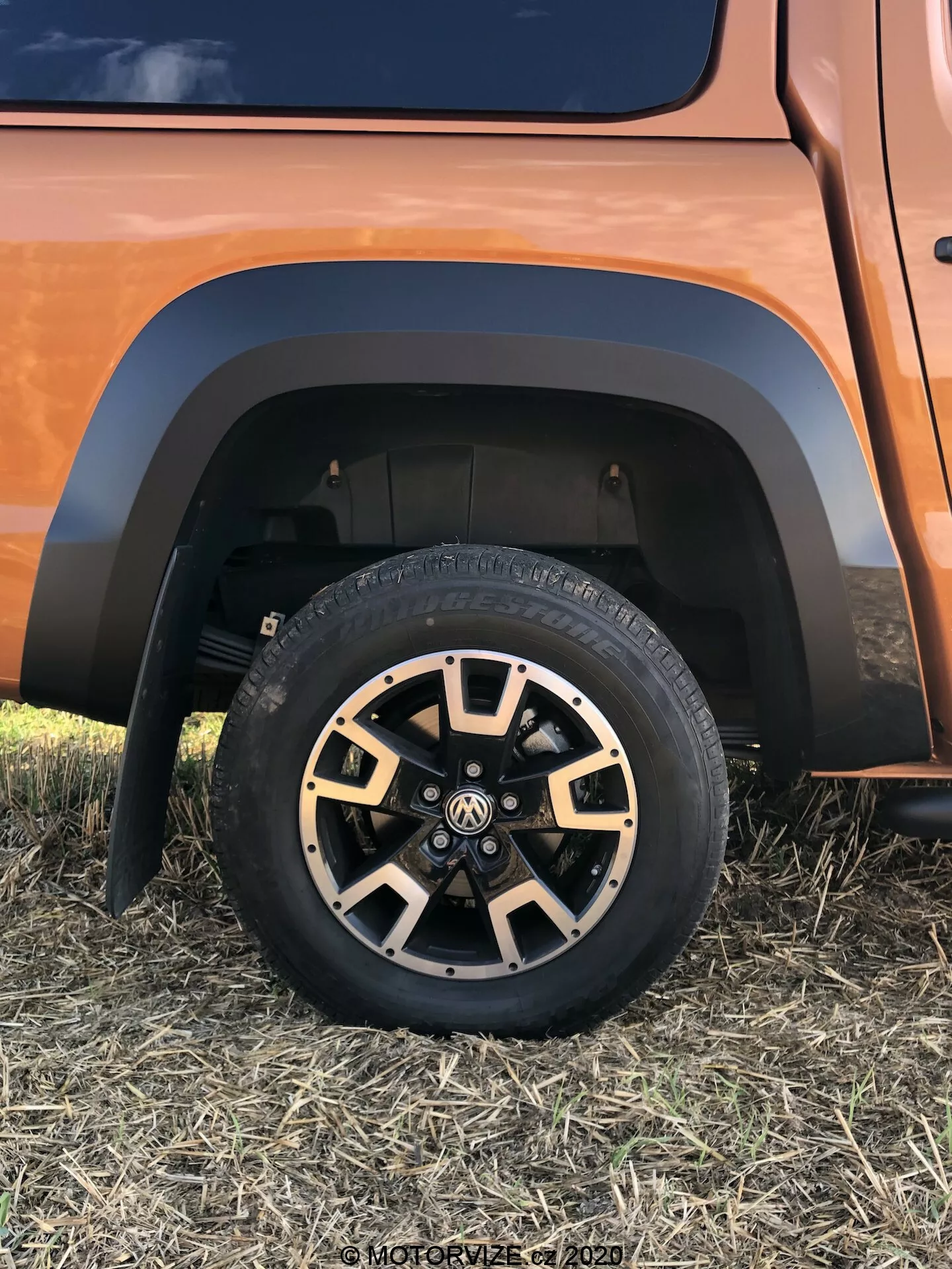 Vista ravvicinata della parte inferiore di un VW Nutzfahrzeuge Amarok 2016, che mostra un robusto pneumatico all-terrain su una ruota in lega multispea con finiture nere e metalliche, un parafango a contrasto e parte del sistema di sospensione in un colore del veicolo terroso o rame, presa da un angolo basso e laterale.