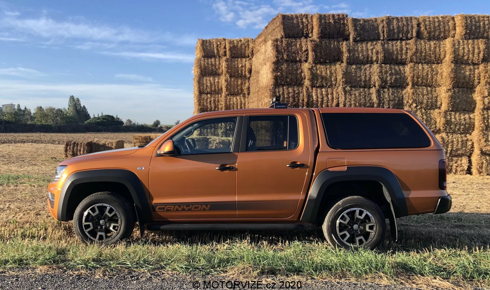 Vista di profilo laterale di un veicolo tipo SUV arancione metallico, probabilmente un SUV di medie dimensioni o un pickup con cabina estesa e copertura del cassone, parcheggiato su terreno erboso piatto con grandi balle di fieno rettangolari sullo sfondo sotto un cielo sereno e soleggiato. Il veicolo presenta linee della carrozzeria audaci, grandi passaruota, finiture nere nella parte inferiore della carrozzeria, ruote in lega robuste, un distintivo visibile nella parte bassa della portiera anteriore e un'antenna sul tetto. La foto mette in evidenza le potenziali capacità fuoristrada e il design senza soluzione di continuità della copertura del cassone.