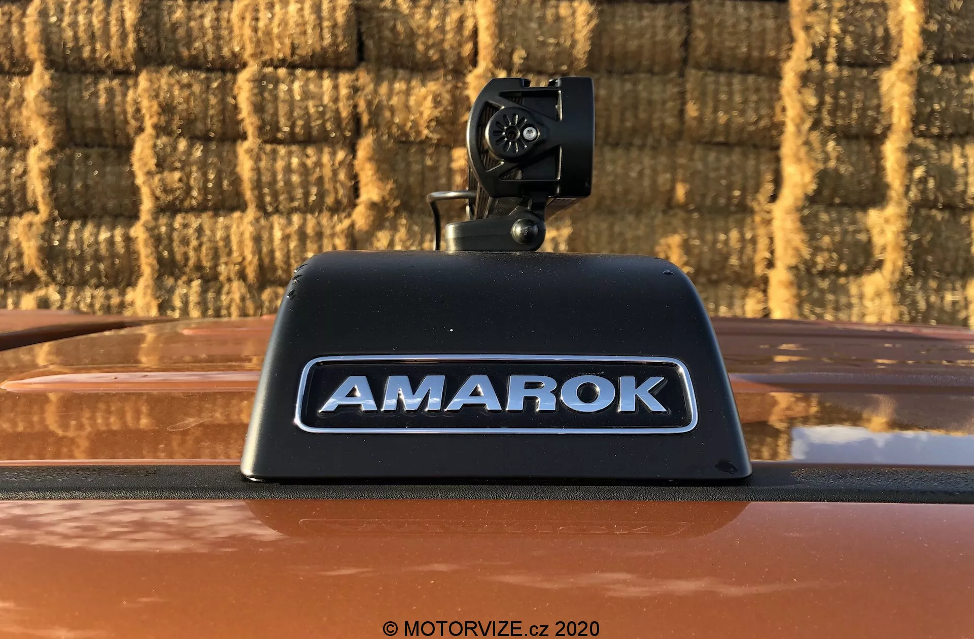 Foto ravvicinata di un pick-up Volkswagen Amarok 2016 che presenta una telecamera di retromarcia aftermarket montata sulla luce di stop alta con 'AMAROK' in lettere in rilievo argentate su una carrozzeria color bronzo metallizzato, all'aperto contro uno sfondo con motivo a mattoni.