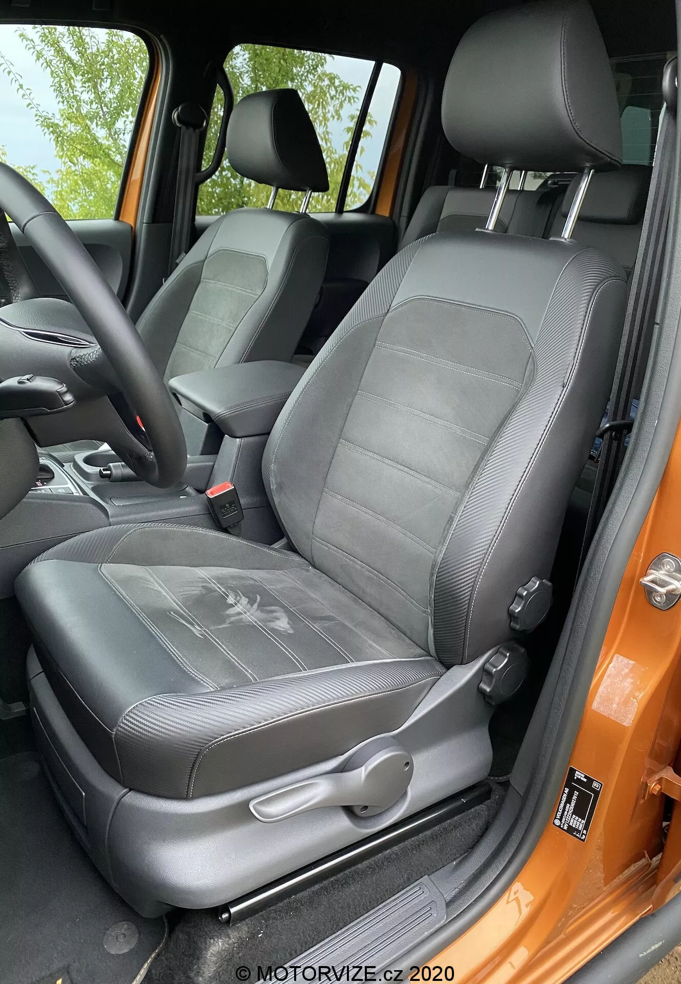Vista ravvicinata del sedile del conducente del VW-nutzfahrzeuge Amarok 2016 dalla prospettiva di chi si trova fuori dalla porta aperta. Il sedile rivestito in similpelle nero presenta un design con texture a coste e un poggiatesta regolabile integrato. Sono visibili i controlli di regolazione manuale e il meccanismo della cintura di sicurezza a tre punti. Il supporto laterale indica un supporto laterale, e il telaio della portiera dipinto in un audace colore arancione contrasta con l'interno nero. Il sedile appare ben mantenuto e illuminato da luce naturale.