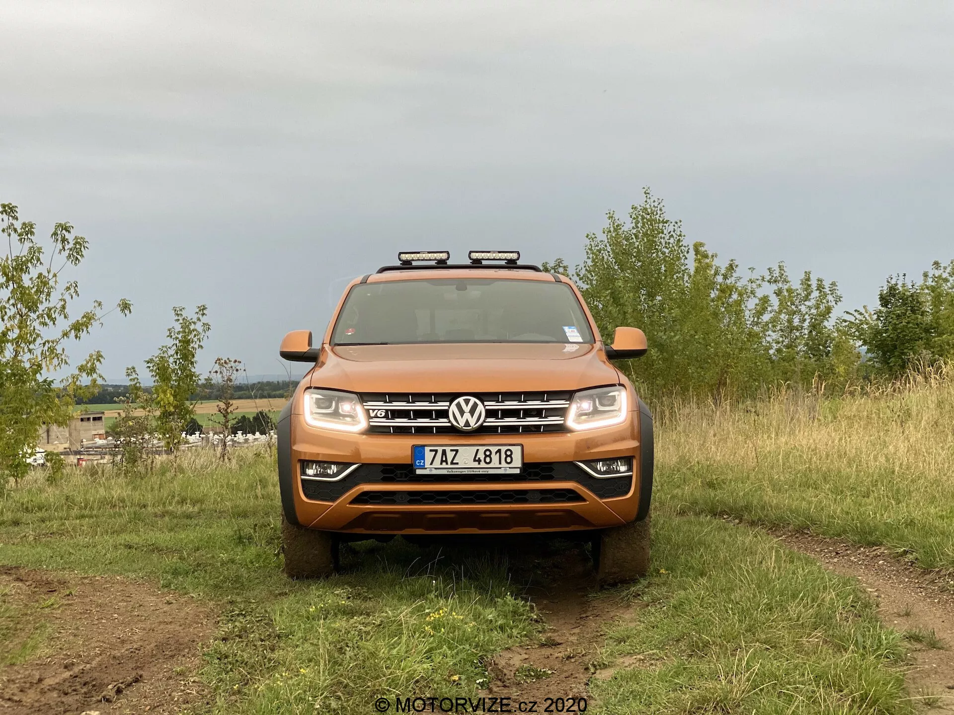 Vista frontale di un SUV Volkswagen Amarok catturato in un contesto all'aperto. Il veicolo arancione presenta il logo Volkswagen in primo piano sulla griglia, listelle orizzontali, fari standard e luci ausiliarie aggiuntive montate sul tetto. L'immagine evidenzia le capacità fuoristrada con pneumatici pesanti parzialmente visibili e sporco sulle superfici del veicolo. È posizionato su un terreno naturale con uno sfondo sfocato, illuminato dalla luce diffusa di un cielo coperto.