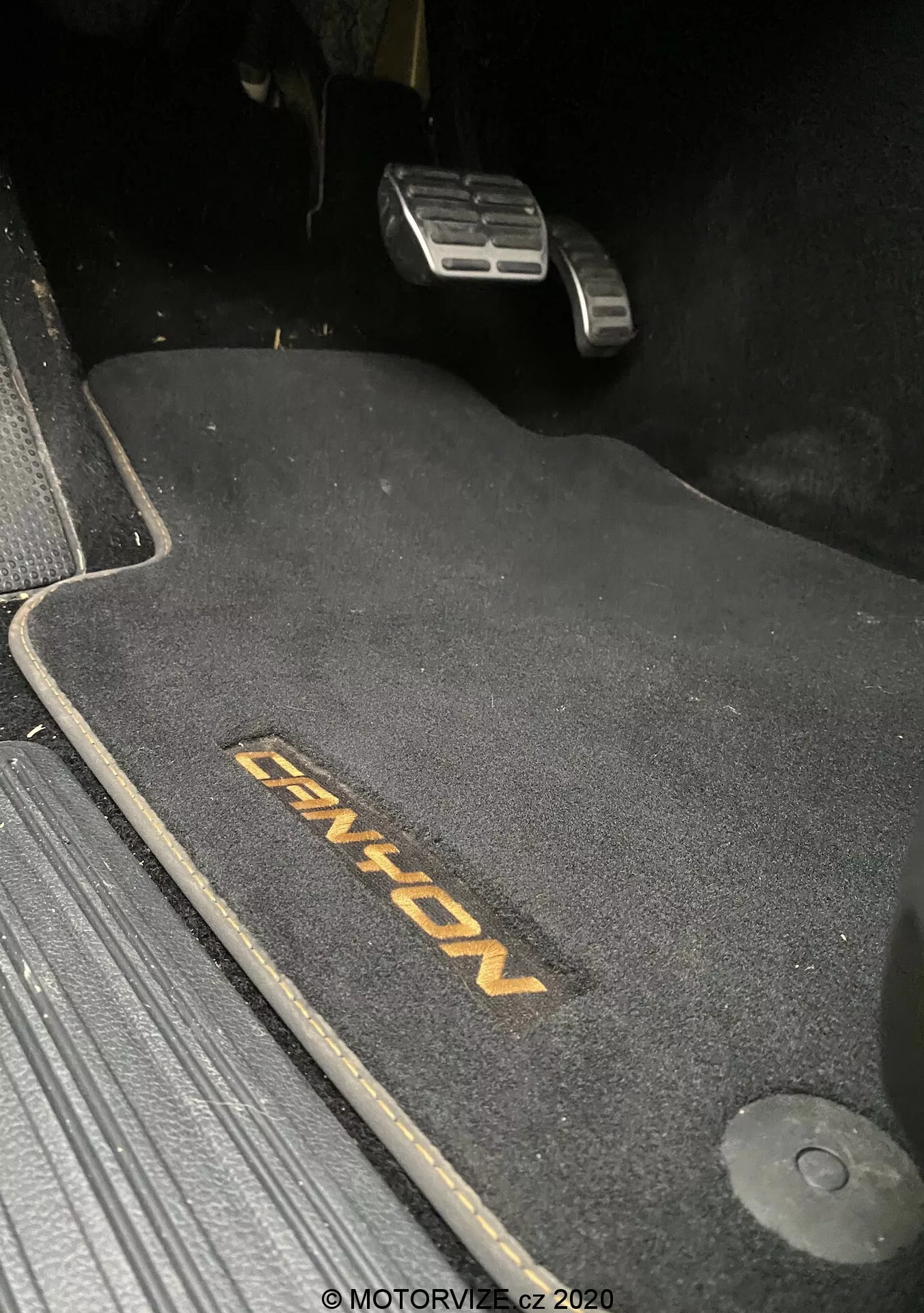 Vista ravvicinata dell'area del pavimento lato guida nel VW-Nutzfahrzeuge Amarok 2016, che mostra un tappetino scuro con ricamo 'ENYON'. Visibili i pedali metallici dell'acceleratore e del freno, con il pedale del freno posizionato più in alto, suggerendo che sia inserito. Il tappetino è sagomato per adattarsi allo spazio, con un gancio di ritenzione sulla destra. Il tappetino contrasta con i dintorni scuri, indicando un interno scuro. La scena è debolmente illuminata, probabilmente scattata di notte o in condizioni di scarsa luminosità.