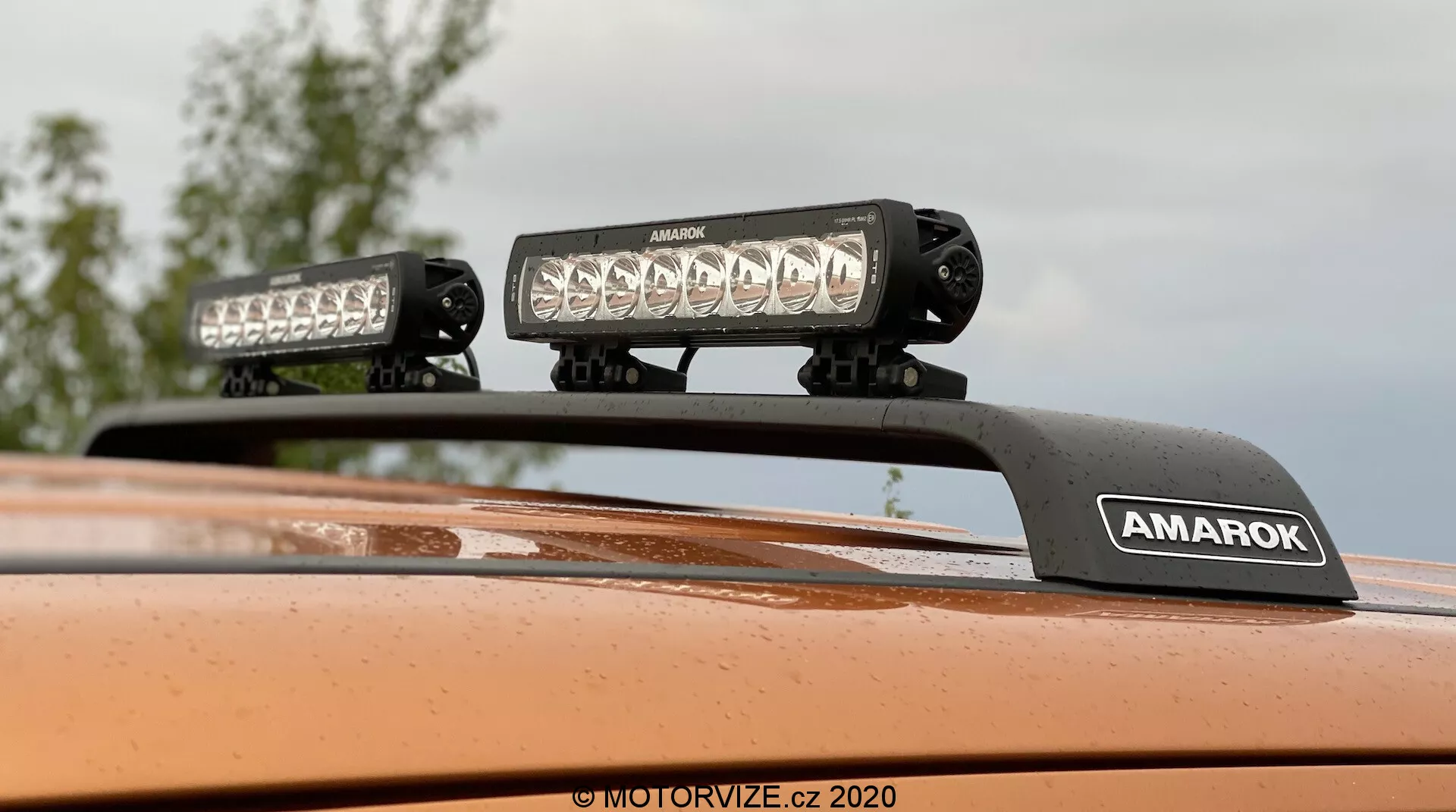 Primo piano verso l'alto del tetto di una Volkswagen Amarok 2016 con equipaggiamento di illuminazione montato sul tetto, due barre luminose LED ad alta intensità su una staffa con marchio 'AMAROK', finitura della vernice in arancione metallizzato; ideale per caratteristiche di veicoli fuoristrada o accessori per veicoli.