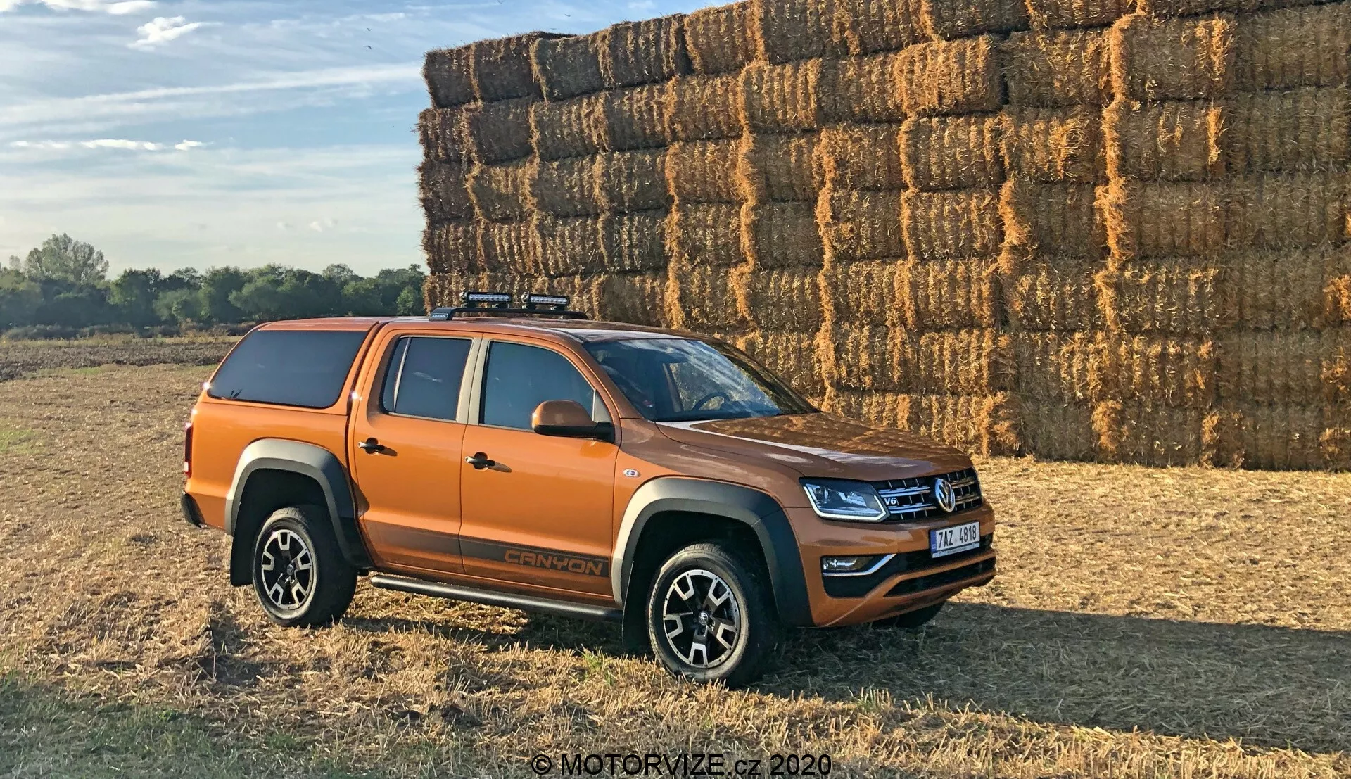 Vista laterale di un pickup Volkswagen Amarok arancione parcheggiato all'aperto su una superficie irregolare, probabilmente di terra o erba. Il veicolo è posizionato a un angolo di 30 gradi rispetto alla fotocamera, mostrando il profilo laterale e una vista parziale della griglia anteriore e dei fari. Il pickup di medie dimensioni moderno presenta linee di design fresche e illuminazione a LED, suggerendo un modello recente. Sono visibili quattro porte che indicano una configurazione a doppia cabina, ruote in lega multiraggio, pneumatici fuoristrada, un copribaule rigido sul cassone della camionetta e un sistema di portapacchi sul tetto. Gli specchietti laterali probabilmente includono indicatori. Fotografo in luce diurna calda con ombre lunghe, un grande mucchio di balle di fieno sullo sfondo suggerisce un ambiente rurale.