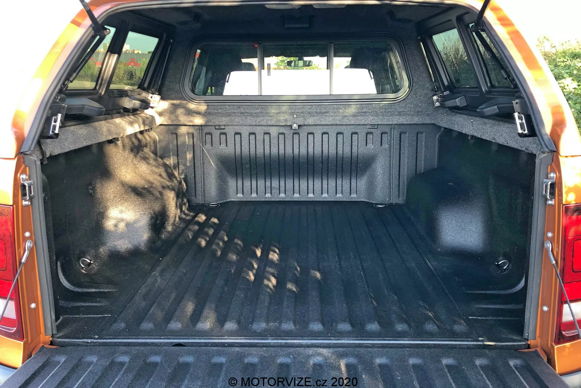 Interno dello spazio di carico del VW Nutzfahrzeuge Amarok 2016, visto da dietro con il portellone aperto, che mostra l'area del bagagliaio. L'area di carico presenta un rivestimento resistente con motivo, pareti laterali rifinite con materiale durevole, punti di ancoraggio per fissare gli oggetti, illuminazione interna per la visibilità, sedili posteriori visibili nella parte anteriore dello spazio e bordo protettivo sul bordo del portellone.