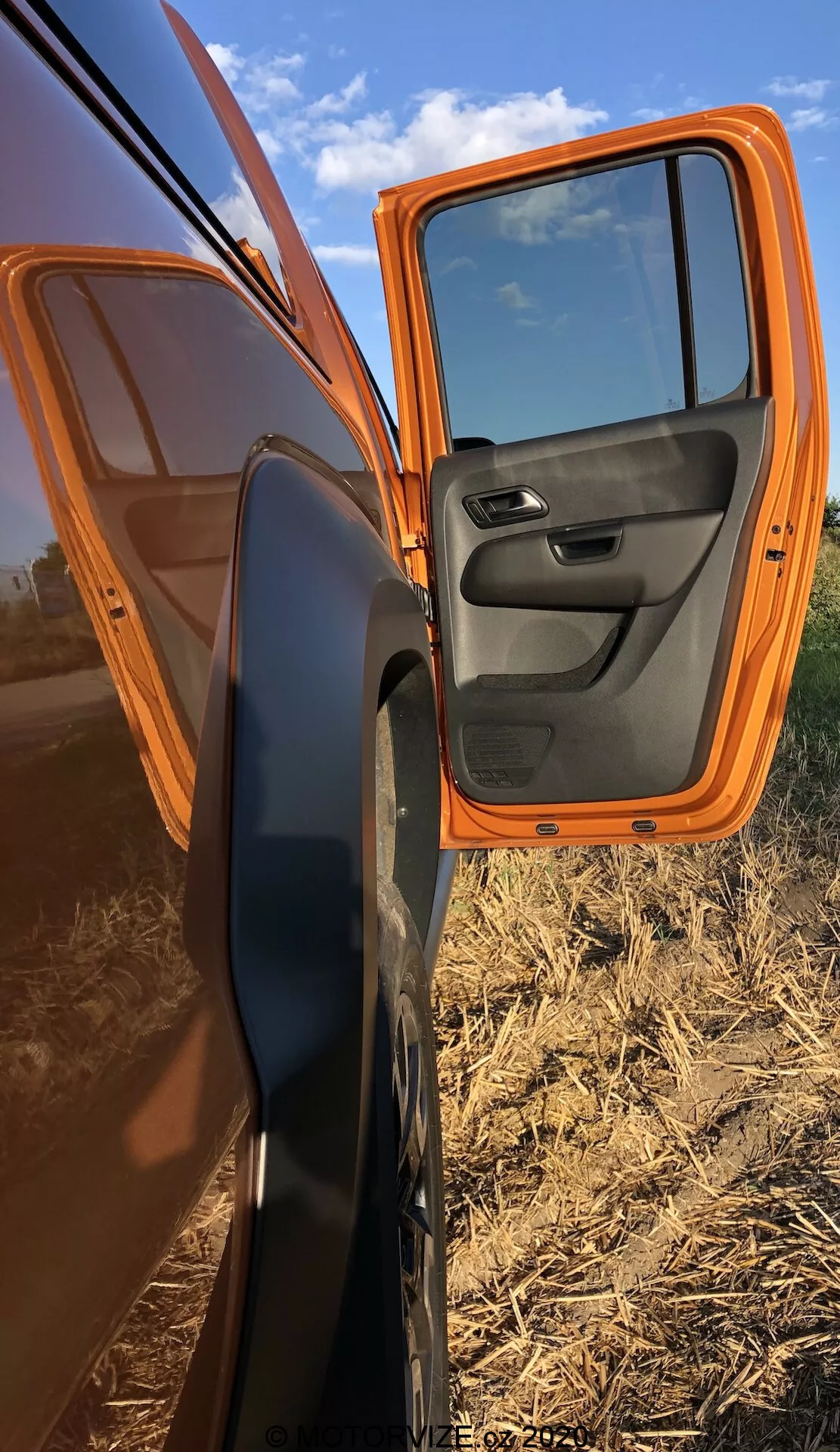 Vista ravvicinata di una porta anteriore del passeggero parzialmente aperta di un VW Nutzfahrzeuge Amarok 2016 arancione, che mostra la vernice esterna arancione brillante e il pannello interno nero con comandi integrati. L'auto è parcheggiata in diagonale, presentando un profilo laterale con la porta aperta ad un angolo di 70 gradi, evidenziando lo specchietto laterale con alloggiamento nero. I riflessi sulla porta mostrano un cielo limpido e terreni agricoli piatti, accentuando l'ambientazione rurale.