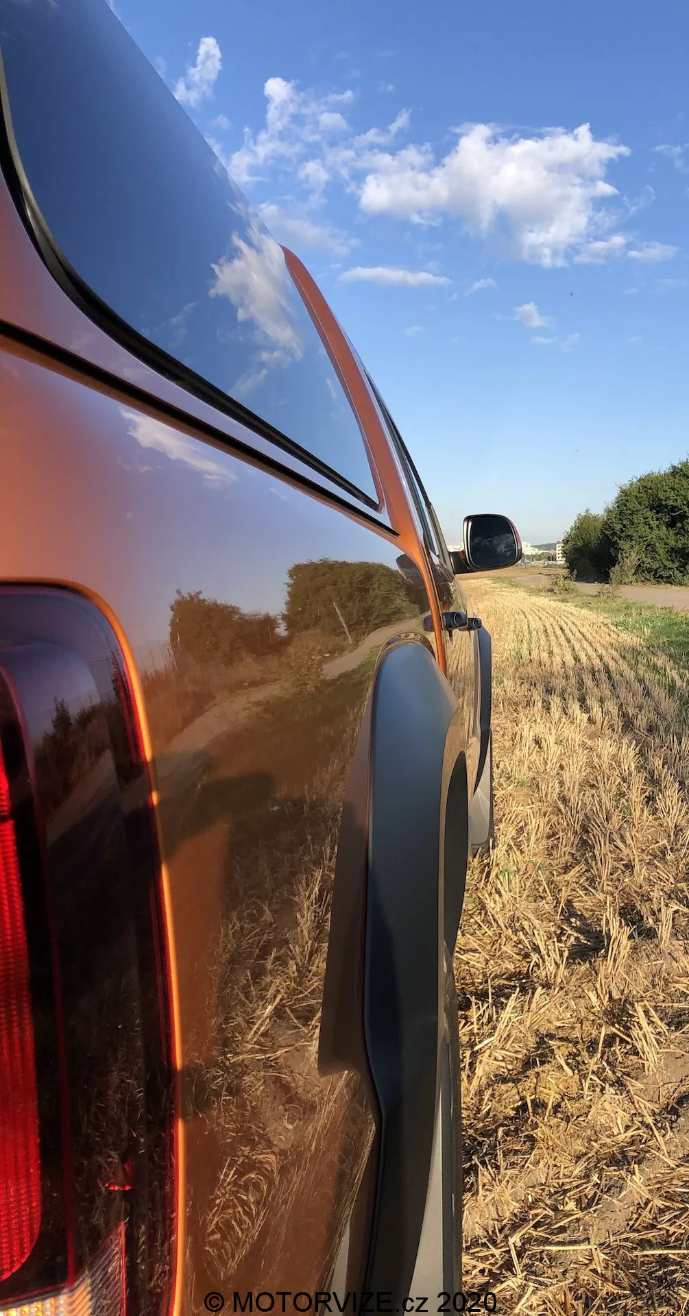 Vista laterale ravvicinata del pannello posteriore destro di un VW Nutzfahrzeuge Amarok 2016, mettendo in evidenza la carrozzeria metallica lucida e riflettente e la luce di coda integrata con lente trasparente. La foto, scattata da un'angolazione bassa, mostra la linea di cintura, il profilo in plastica nera e il paesaggio rurale del campo raccolto con un cielo azzurro.
