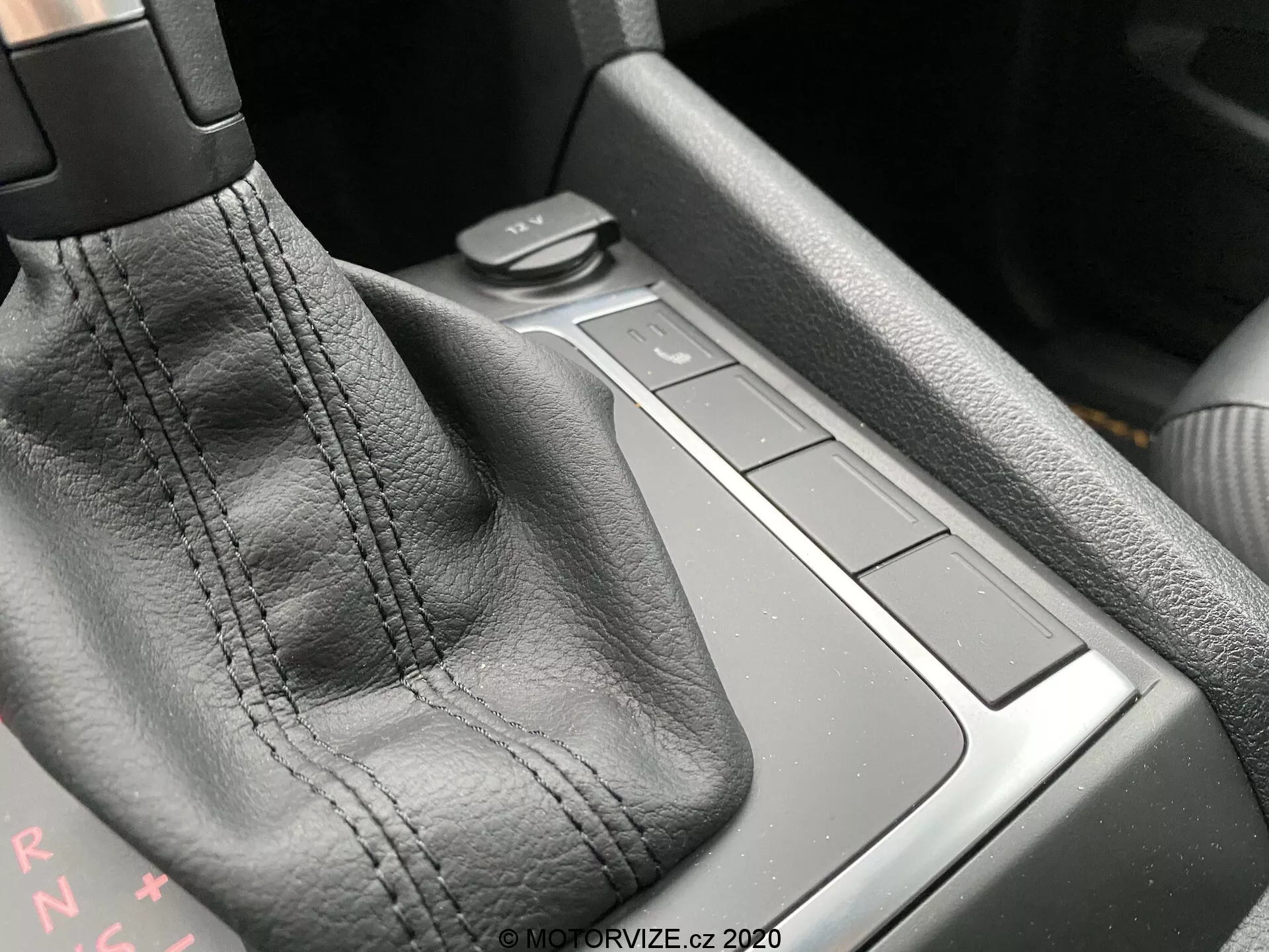 Immagine ravvicinata della console centrale di un VW Nutzfahrzeuge Amarok 2016, focalizzata sulla zona del cambio. La leva del cambio, rivestita in pelle nera con cuciture visibili, è l'elemento principale. Le lettere 'PRNDS' dietro la leva indicano una trasmissione automatica. A destra ci sono quattro pulsanti quadrati a filo con la superficie della console, probabilmente per controllare varie funzioni dell'auto. L'area dietro i pulsanti potrebbe essere un piccolo vano portaoggetti. I materiali e la qualità costruttiva suggeriscono un alto livello di artigianato. La luce ambientale proveniente dall'angolo in alto a sinistra mette in risalto i dettagli.