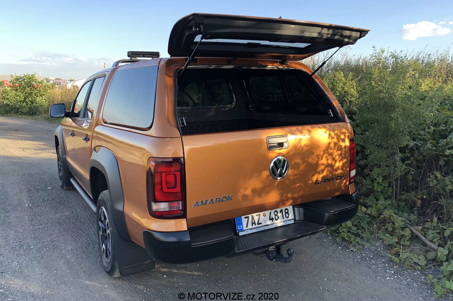 Vista posteriore tre quarti di un Volkswagen Amarok 2016 in arancione metallico, con enfasi sul coperchio del letto aperto, il gancio di traino, le porte posteriori della doppia cabina e parte della ruota posteriore destra e del paraurti, ambientato in un contesto rurale al tramonto. Foto di MOTORVIZE.cz, 2020.