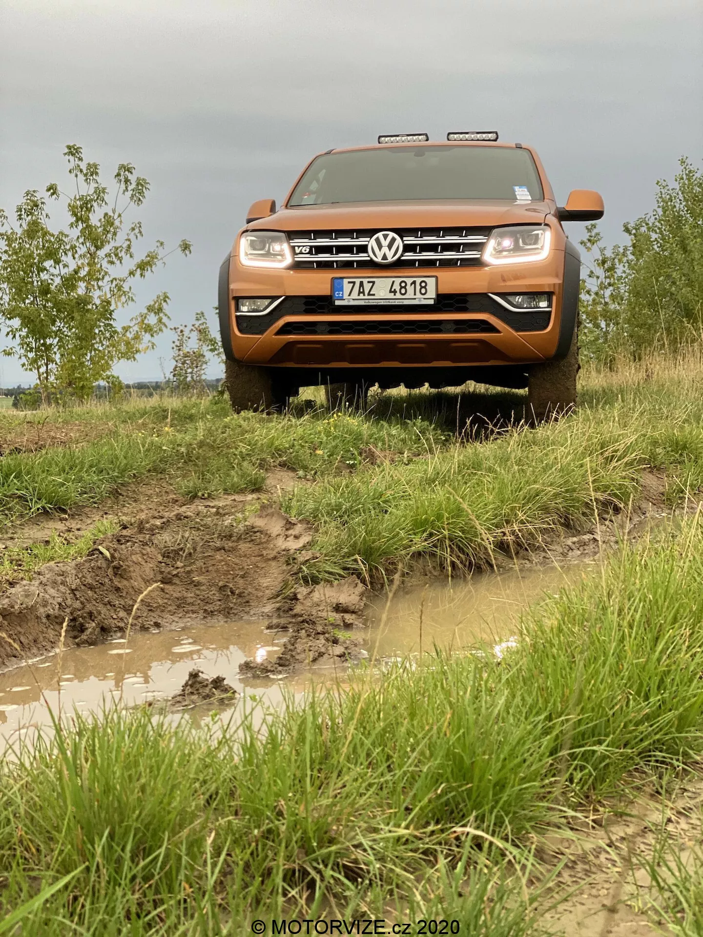 Vista frontale di un veicolo fuoristrada Volkswagen Amarok del 2016 color arancione, evidenziando la griglia anteriore, il paraurti, il cofano e l'assemblaggio dei fari. Fotografato da un angolo basso per enfatizzare la sua posizione elevata, l'immagine rivela un sentiero accidentato e fangoso con pozze d'acqua e un esterno schizzato di fango, accentuando le sue capacità fuoristrada. Il veicolo presenta l'imponente griglia Volkswagen con emblema, fari rettangolari e luci ausiliarie sopra la griglia. La targa è visibile sul robusto paraurti anteriore, mentre il cofano piatto e allungato sottolinea la dimensione e la potenza dell'Amarok.