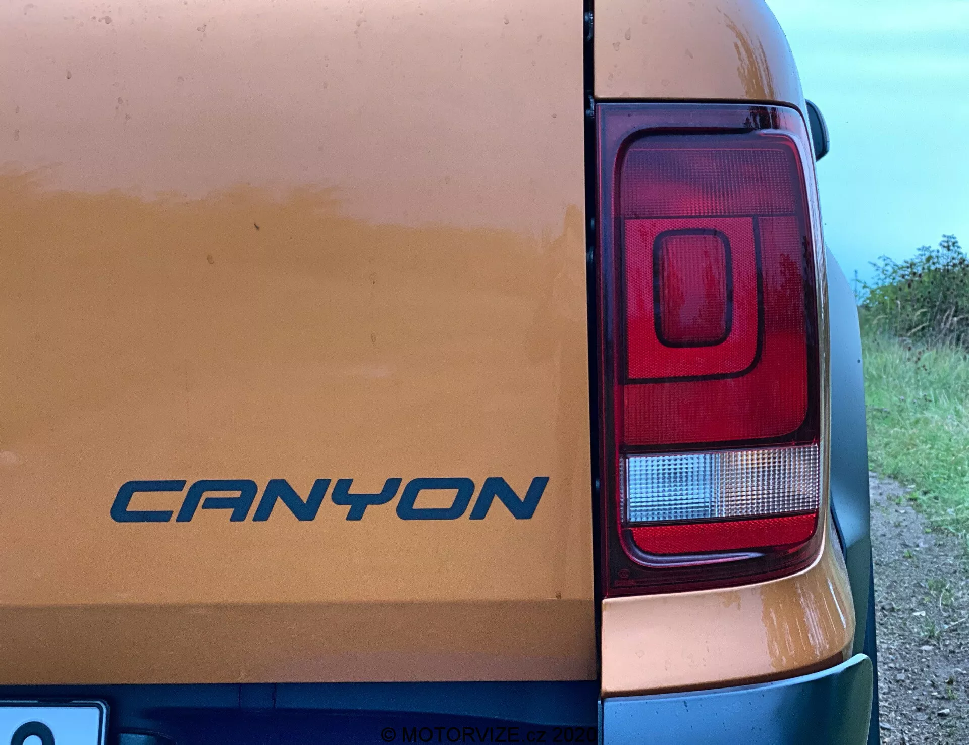 Vista ravvicinata dell'angolo posteriore destro di un veicolo VW Nutzfahrzeuge Amarok 2016. Il badge 'CANYON' è visibile sulla porta posteriore in mezzo a segni di lieve sporcizia, suggerendo un uso su strade sterrate. La fotografia evidenzia principalmente il cluster del fanale posteriore rettangolare e moderno con sezioni per il freno, la retromarcia e probabilmente le frecce direzionali. Scattata da un angolo laterale di circa 45 gradi, l'immagine cattura parzialmente la carrozzeria del veicolo con uno sfondo rurale o semi-rurale.