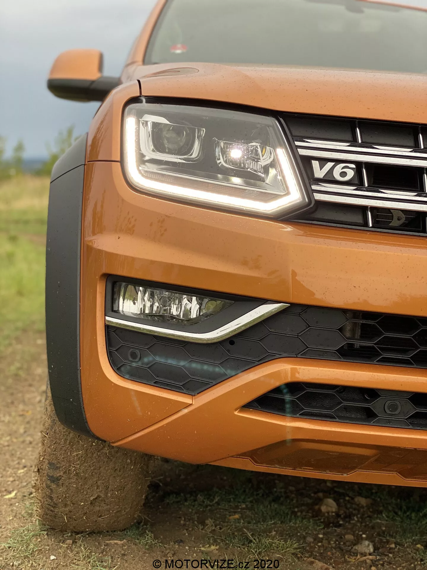 Primo piano dell'angolo anteriore sinistro di una VW Nutzfahrzeuge Amarok arancione del 2016 che mostra il faro con lente chiara moderna e tecnologia a LED, fari fendinebbia con cornice nera, griglia parzialmente visibile con badge V6, paraurti anteriore con rete a nido d'ape nera, design del cofano sagomato e ruota parzialmente visibile, con sfondo sfocato che enfatizza la texture e il design robusto dell'auto