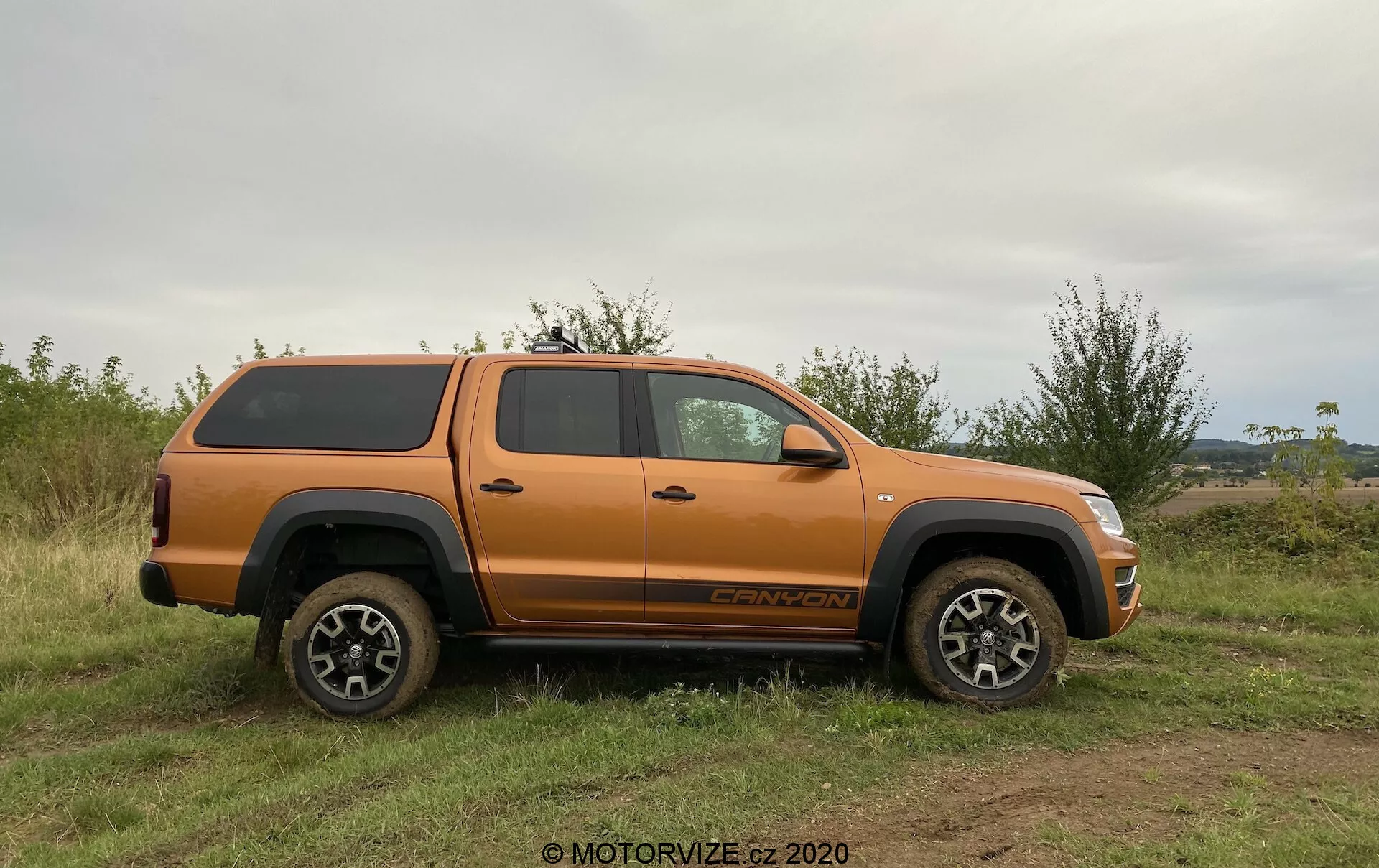 Profilo laterale di un pick-up VW Nutzfahrzeuge Amarok 2016 arancione con una copertura rigida sulla cassone, parcheggiato su terreno erboso e irregolare. Il veicolo presenta un'elevata altezza dal suolo, ruote e pneumatici robusti, e un design in lega multi-razze adatto per uso fuoristrada. Elementi visibili includono specchietti laterali, maniglie delle portiere, parafanghi, pedane, cabina spaziosa a quattro porte, badge sulla ruota anteriore e testo del livello di allestimento sopra la ruota posteriore. La luce naturale e l'angolo equilibrato mettono in risalto i dettagli del veicolo senza ombre pronunciate, enfatizzando le capacità fuoristrada e lo stoccaggio sicuro del carico.