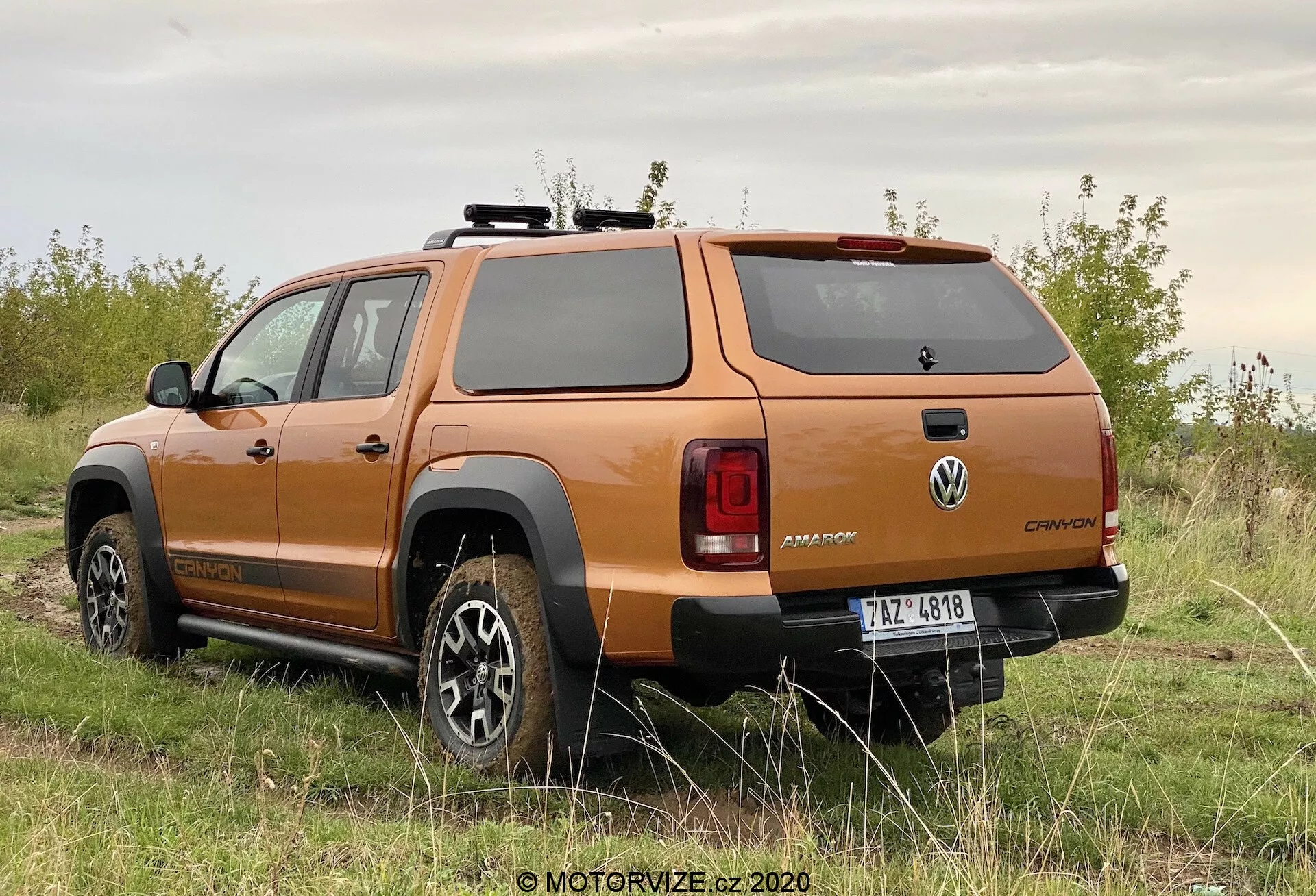 Vista posteriore tre quarti di un pick-up arancione Volkswagen Amarok 2016, che mostra il design delle ruote, il profilo laterale, il finestrino posteriore, i fanali posteriori, il robusto paraurti posteriore e la copertura rigida del letto, parcheggiato su una superficie non asfaltata con vegetazione, evidenziando le sue capacità fuoristrada.