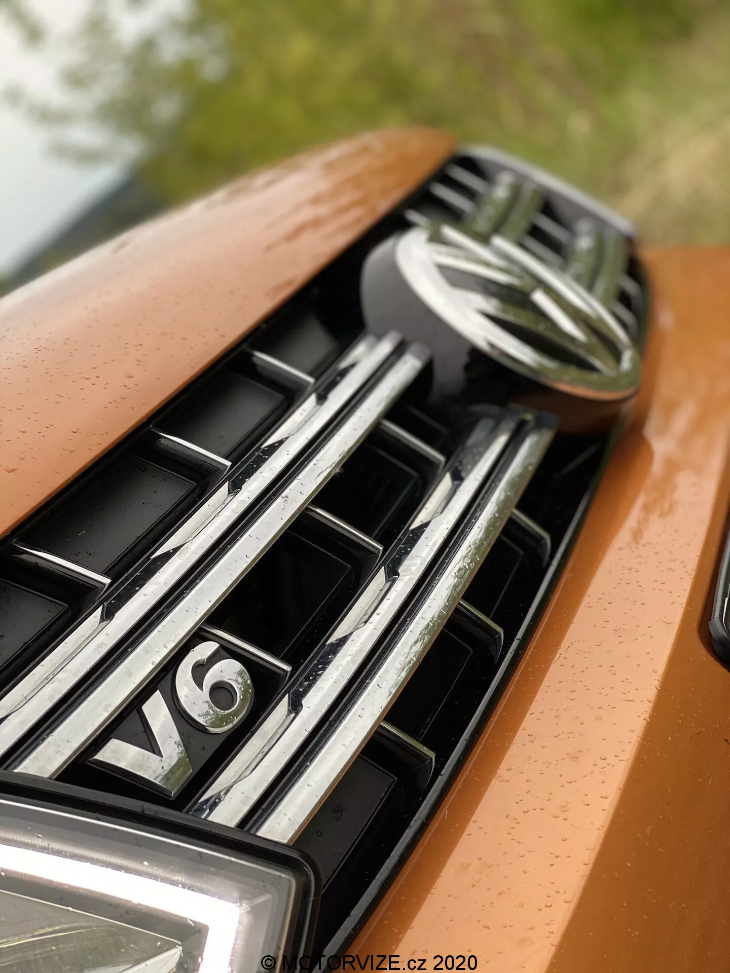 Primo piano del cofano e della griglia di un'auto, focalizzato sulla parte superiore che mostra un emblema centrale e un badge del motore V6. La griglia presenta lamelle orizzontali riflettenti in cromo, indicative di un veicolo di lusso. La verniciatura dell'auto è di un colore arancione o bronzo metallizzato con un'illuminazione morbida e diffusa che accentua il design fluido e l'aspetto premium.