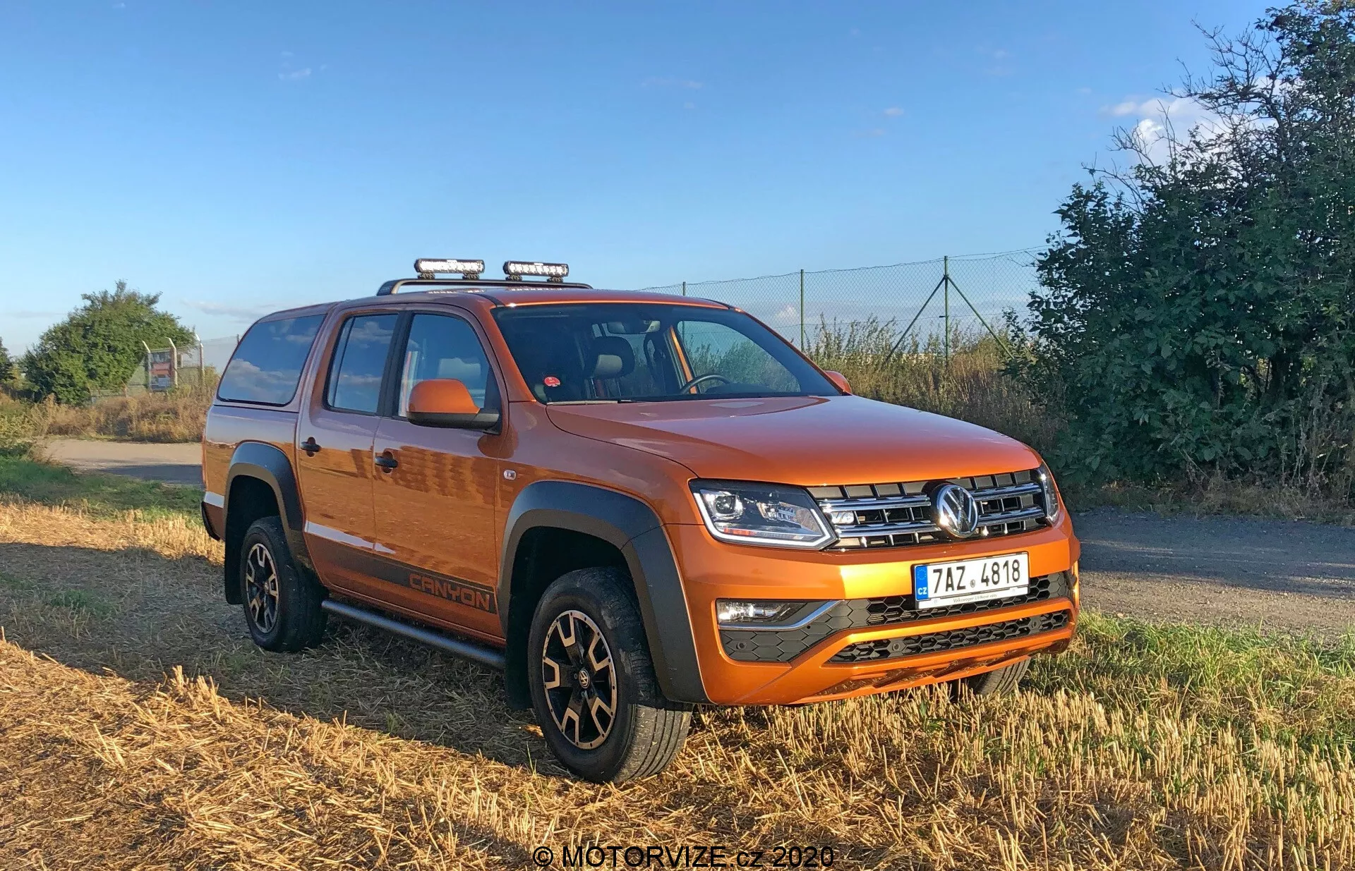 Immagine di un vivace pick-up a quattro porte VW Nutzfahrzeuge Amarok 2016 arancione scattata con un'angolazione di tre quarti, che mostra la parte anteriore e laterale del veicolo. L'immagine mette in evidenza la griglia frontale orizzontale, i complessi fari a LED, le luci ausiliarie sul tetto, il robusto paraurti con fendinebbia, la piastra paramotore, la sezione centrale rialzata del cofano, protezioni nere sugli archi delle ruote, cerchi neri con pneumatici multiuso, gradini laterali, specchietti con indicatori di direzione integrati, cassone coperto con tonneau e binari sul tetto per una maggiore capacità di stoccaggio.