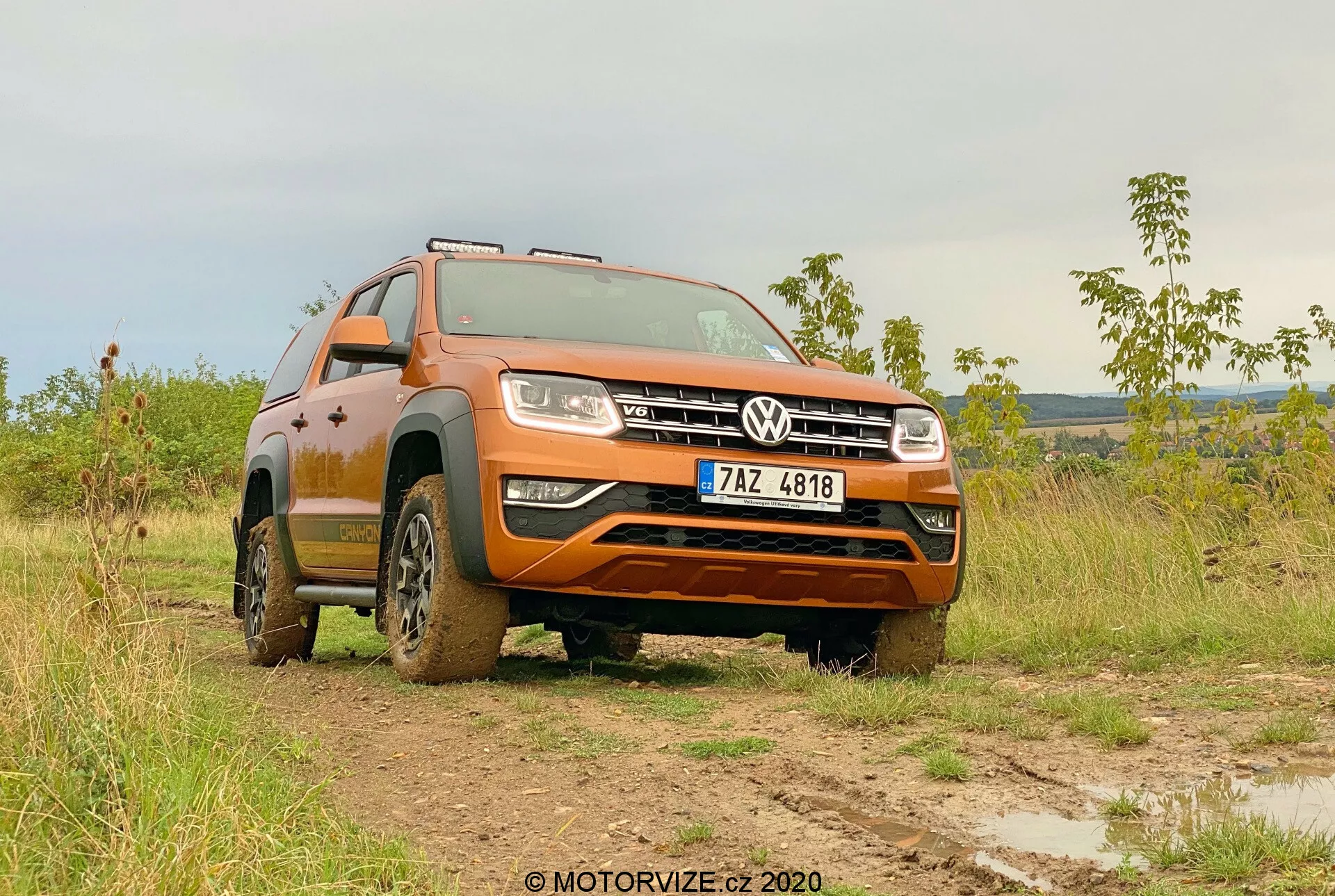 Angolo anteriore di tre quarti basso di un Volkswagen Amarok 2016 in un campo fangoso, che mostra la sua verniciatura arancione, la griglia anteriore con il badge Volkswagen, i fari, il design delle ruote e la capacità fuoristrada. La robustezza del veicolo è evidenziata dal fango sui pneumatici e sui lati inferiori, con le ruote anteriori girate a sinistra che enfatizzano i disegni del battistrada e l'altezza di guida. Ambientato in uno sfondo con luce soffusa, sia di mattina che di tardo pomeriggio, l'immagine sottolinea la versatilità del Amarok in tutti i terreni e il suo avanzato sistema di illuminazione. La targa è visibile e deve essere oscurata per la privacy.