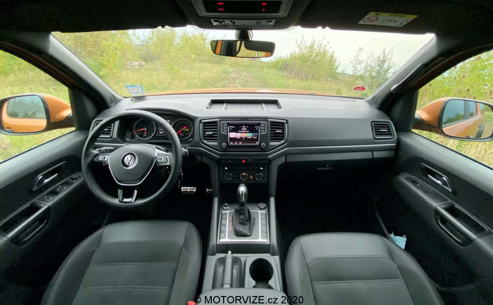 Vista dell'abitacolo anteriore del VW-Nutzfahrzeuge Amarok 2016. Scattata dalla prospettiva del sedile posteriore, questa immagine mostra il volante centrale con comandi integrati, il cruscotto analogico con un piccolo display digitale, il sistema di infotainment al centro del cruscotto con comandi audio e clima, la leva del cambio che indica la trasmissione automatica e sedili in pelle. L'immagine cattura anche lo specchietto retrovisore, gli specchietti retrovisori esterni e i finestrini laterali anteriori, evidenziando caratteristiche tecnologiche e di comfort.