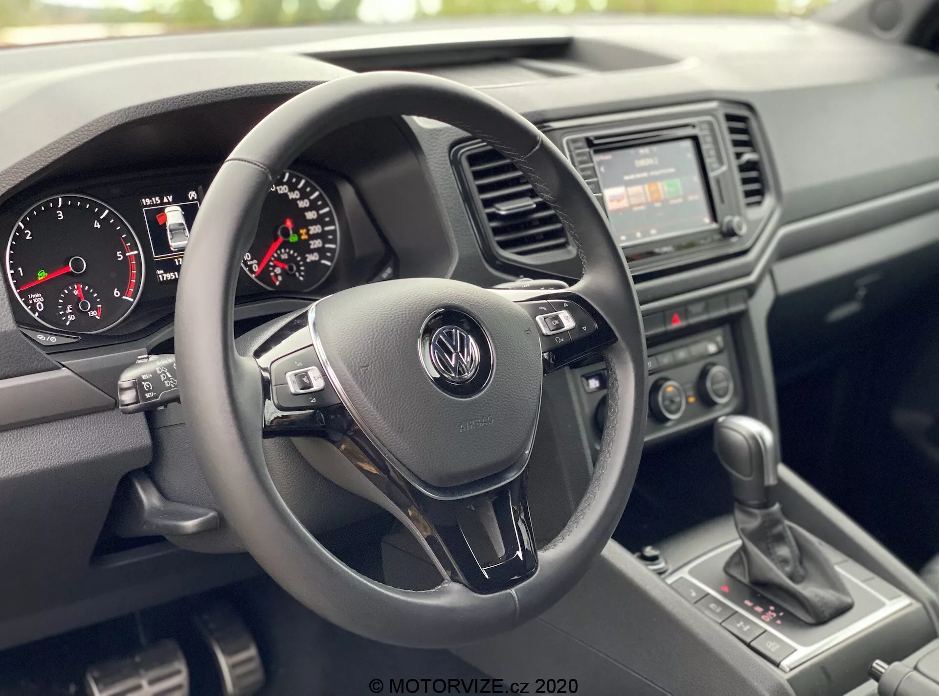 Vista interna del VW Nutzfahrzeuge Amarok 2016 dalla prospettiva del passeggero anteriore, che mostra l'area del posto di guida. Focus centrale su un volante a tre razze con logo Volkswagen e pulsanti di controllo multifunzione, quadro strumenti con tachimetro analogico, contagiri e piccolo display digitale, radio di base con interruttori di controllo del clima manuale sulla console centrale, leva del cambio manuale e materiali plastici texturizzati per cruscotto e porte, evidenziando un design durevole e pratico.