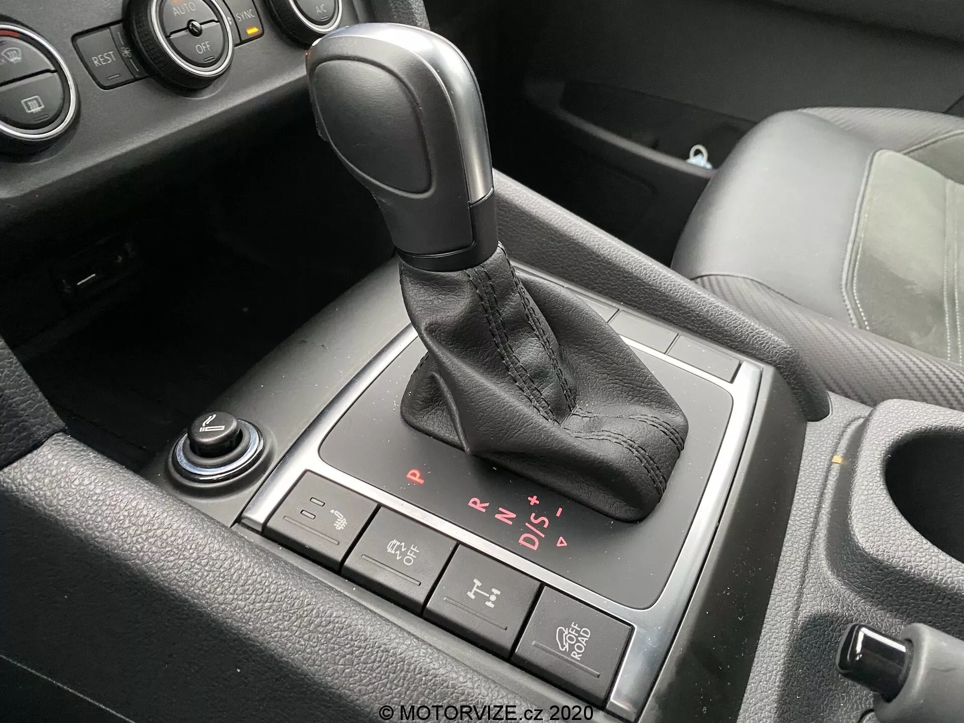 Vista dettagliata dell'area del cambio nell'interno del VW Nutzfahrzeuge Amarok 2016. Il cambio automatico con impugnatura nera e cuffia in pelle è posizionato centralmente nell'immagine con visibili opzioni P, R, N, D e modalità manuale sequenziale. Dietro la leva del cambio, c'è un pulsante del freno di stazionamento elettronico, mentre una manopola rotante parzialmente visibile e un'altra manopola sono situate davanti e in basso a sinistra, rispettivamente. Sono visibili anche viste parziali del portabicchieri e della console centrale con accenti in plastica nera e alluminio o cromo, che indicano un veicolo moderno con finiture interne di livello medio o superiore.