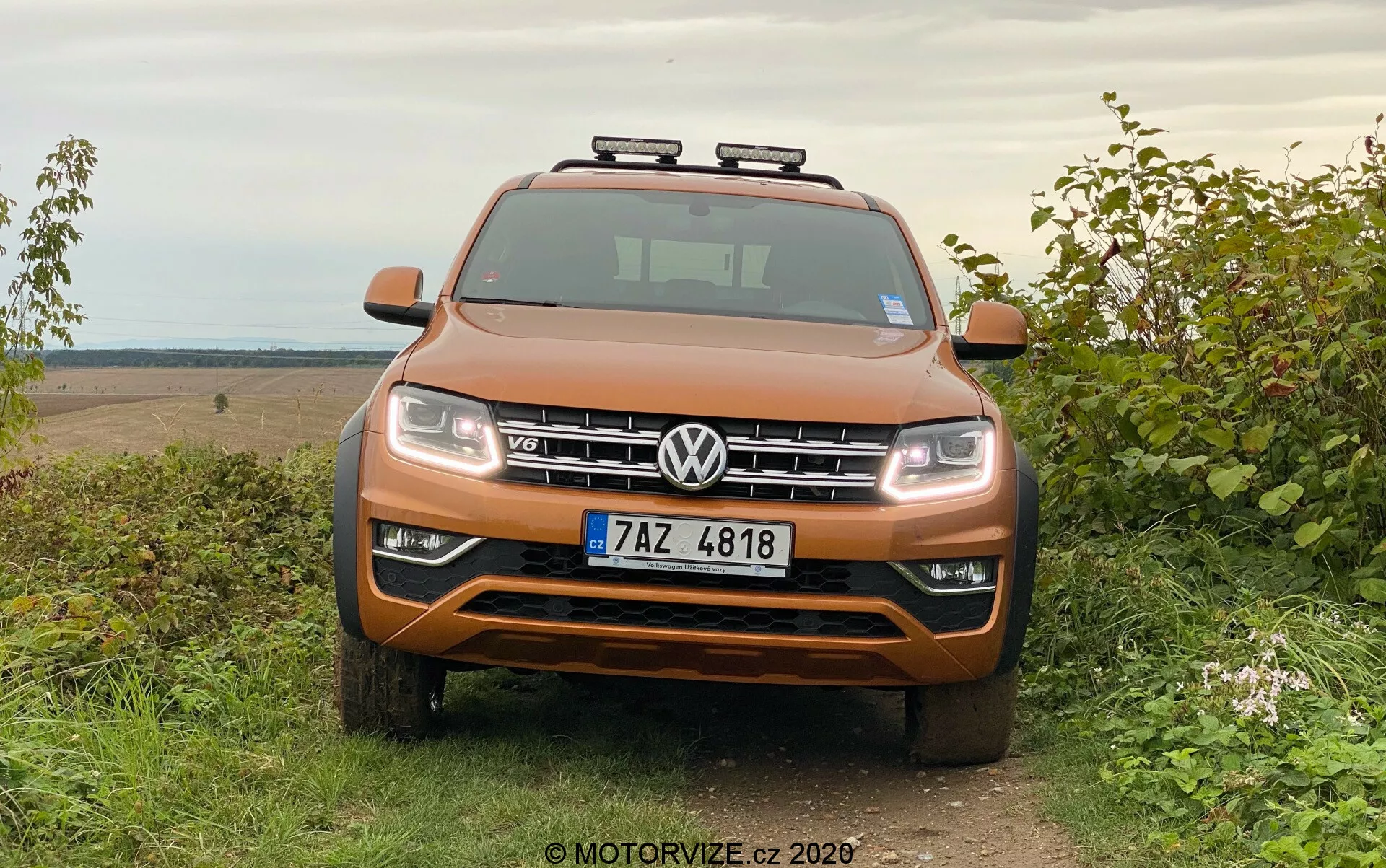 Vista frontale di un SUV Volkswagen Amarok arancione metallizzato parcheggiato sull'erba con sfondo rurale all-terrain, che mostra griglia simmetrica, fari, accenti cromati e luci ausiliarie sul tetto.